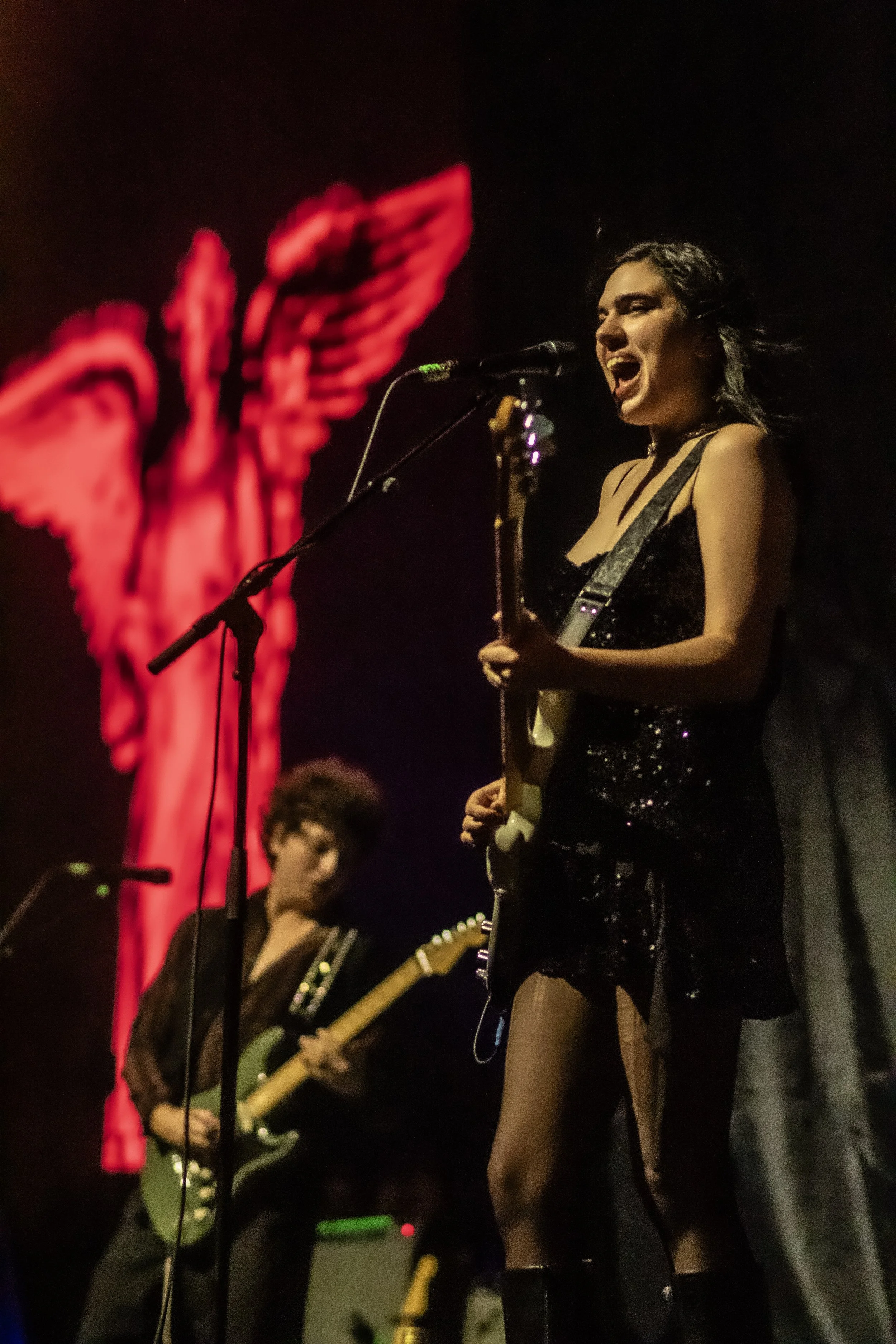 A female musician singing and playing an electric guitar on stage, with a red neon sign in the shape of wings in the background, and another guitarist playing in the background.