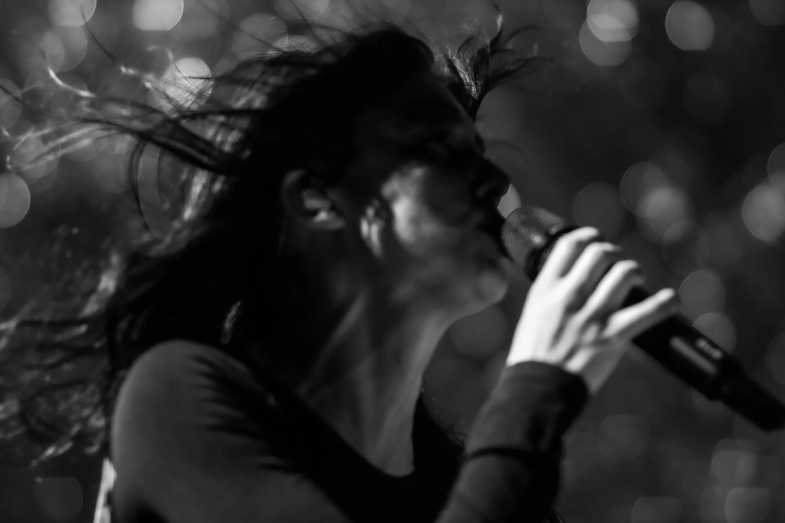 A woman with windswept hair singing into a microphone in black and white.