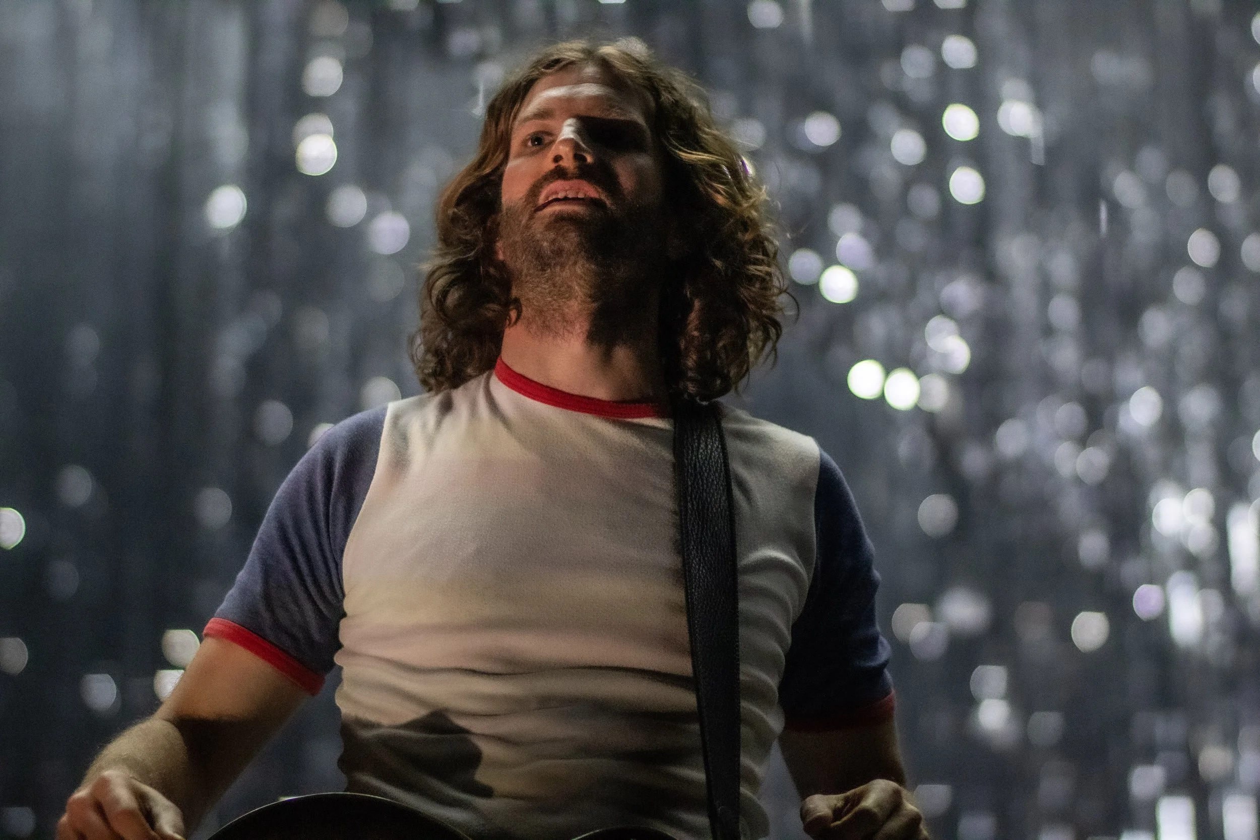 A man with long curly hair and a beard, wearing a vintage baseball T-shirt, standing in a forest with bokeh lights in the background, looking upward with a serious expression.
