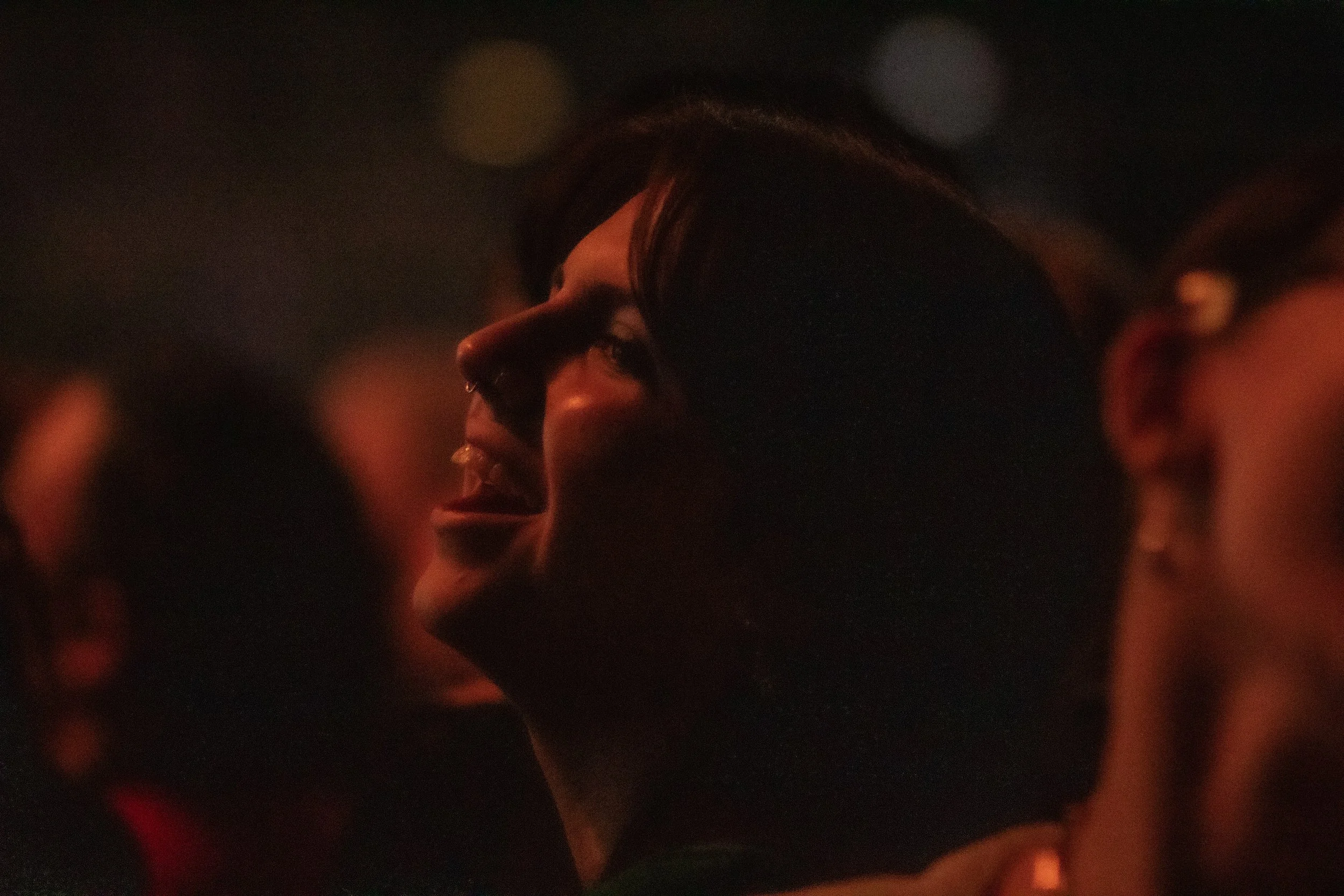 A woman with dark hair and a nose piercing is smiling and looking at someone or something, illuminated slightly by warm lighting, in a dark environment with two other out-of-focus people nearby.