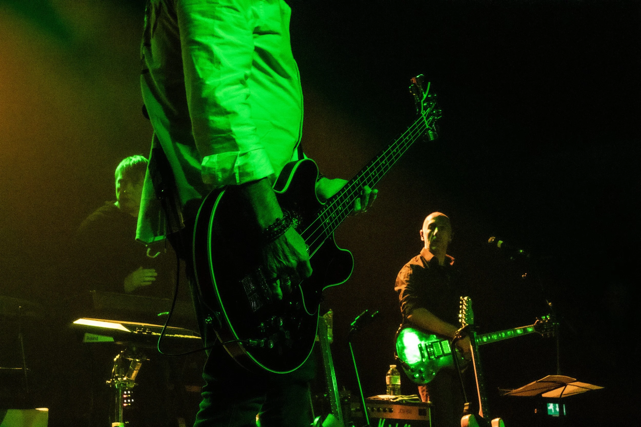 Musicians performing on stage with green lighting, one playing an electric guitar and another with a microphone, with a keyboard in the background.