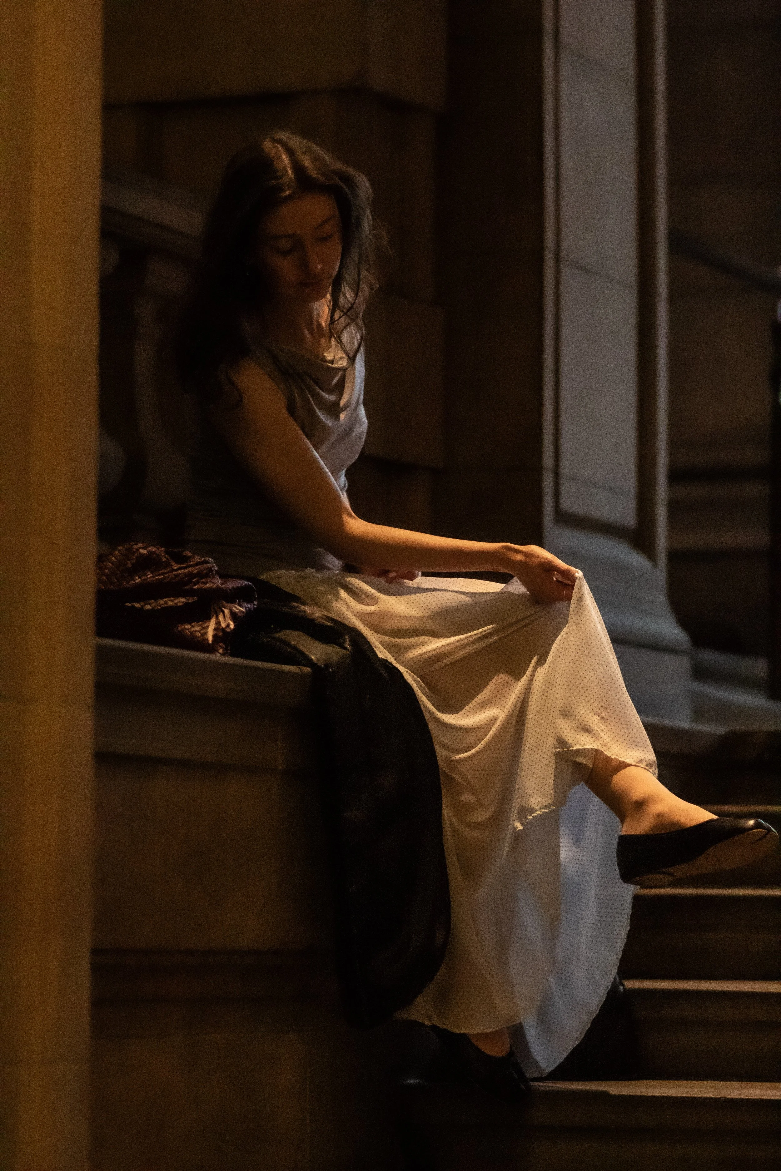 A woman sitting on a bench in a dimly lit indoor setting, adjusting her long, light-colored dress.