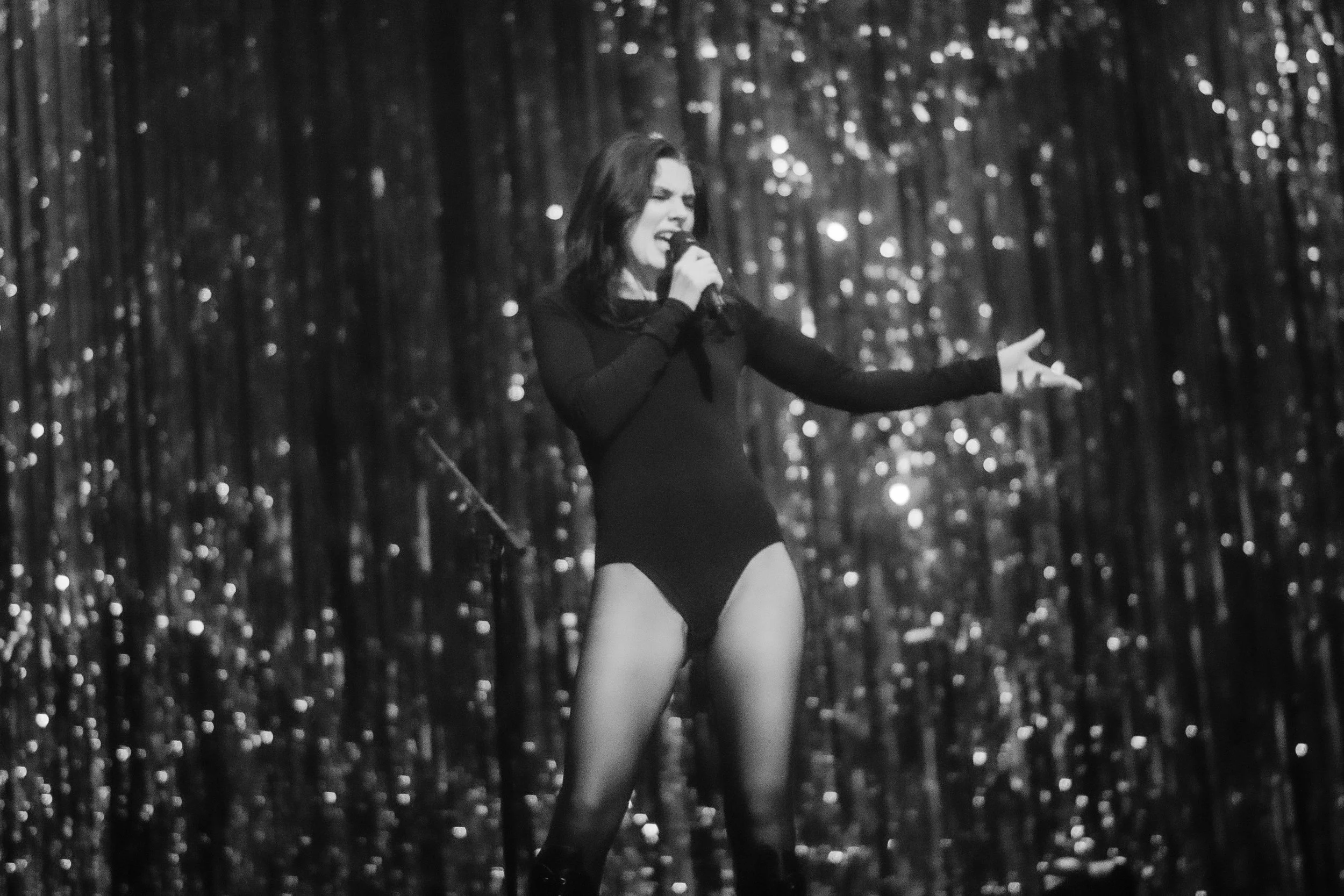 A woman performing on stage, singing into a microphone, wearing a black outfit and tights, with a glittery backdrop.