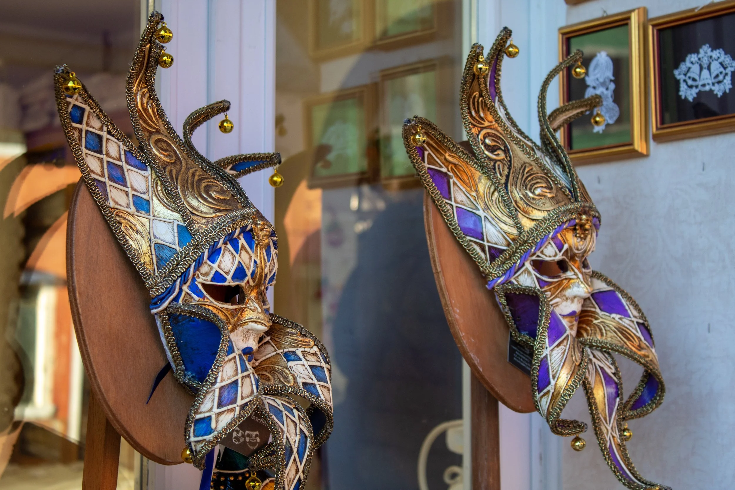 Two ornate carnival masks hanging on a wall, decorated with gold, blue, purple, and white details, featuring intricate patterns and jester-like extensions.