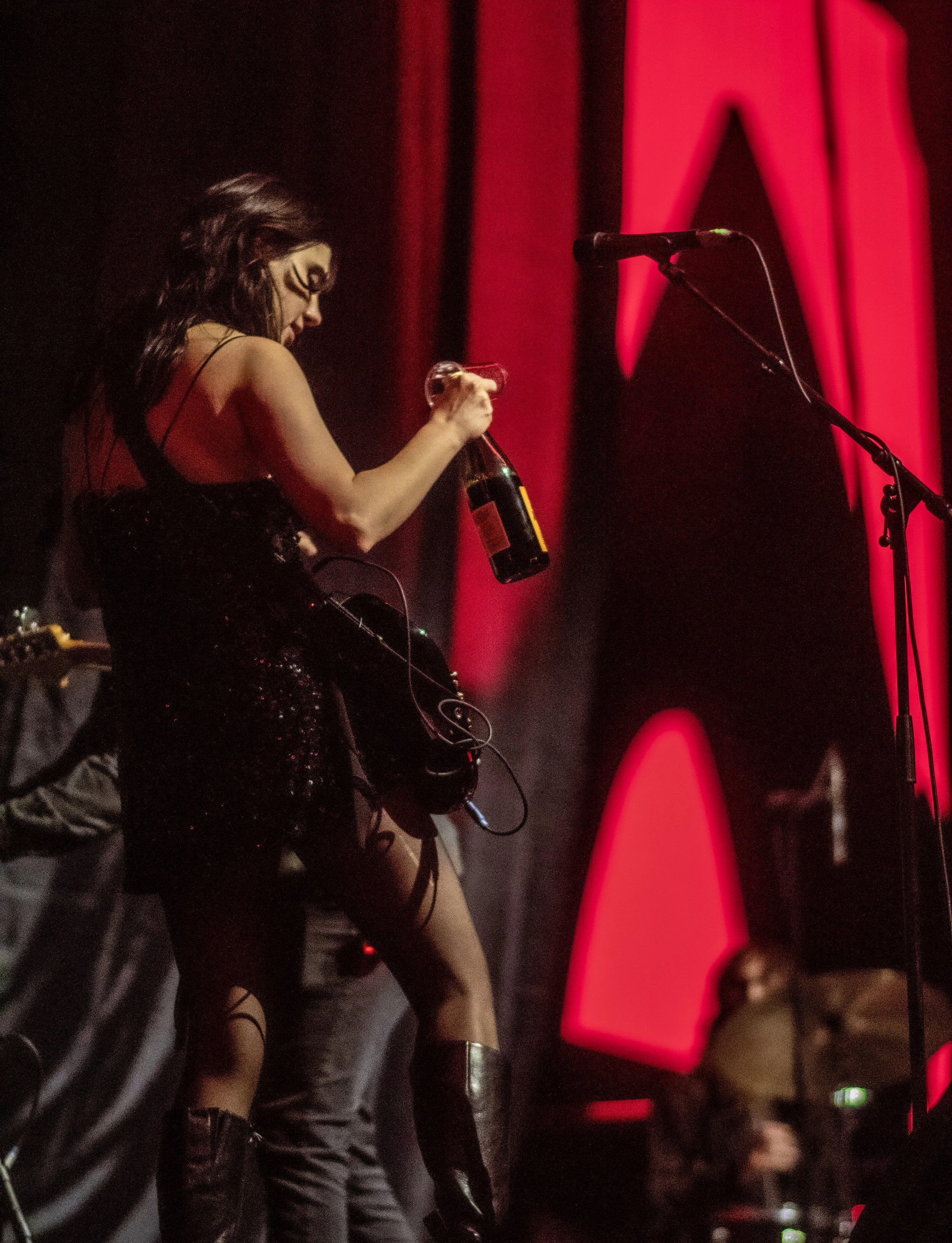 A woman on stage holding a beer bottle, wearing a black outfit with thigh-high boots, with a red backdrop and a microphone stand nearby, and a drummer visible in the background.