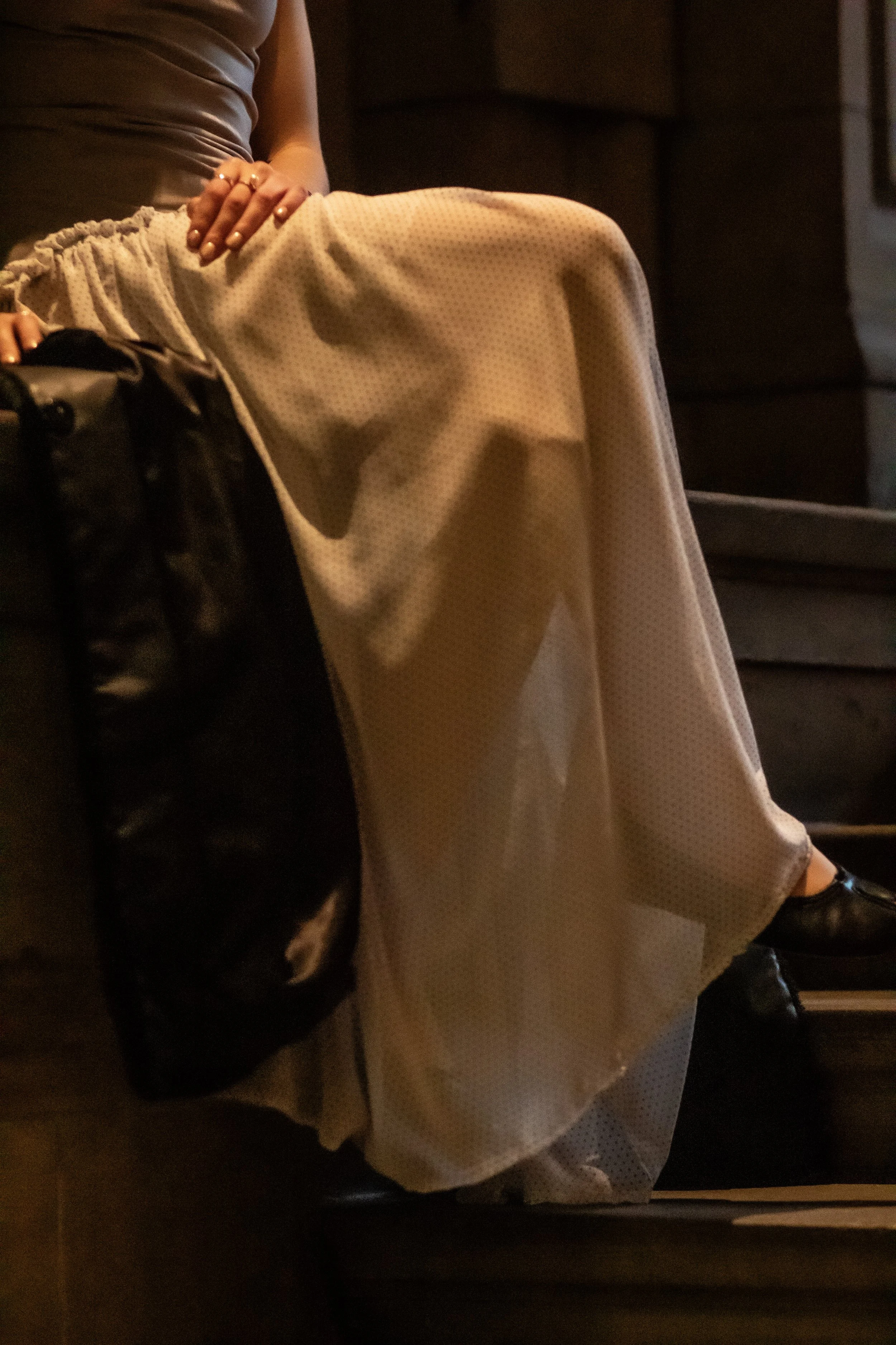 A person sitting in a dimly lit environment, wearing beige wide-leg pants, a grey top, and black shoes. They are resting one hand on their lap, with the other hand not visible.