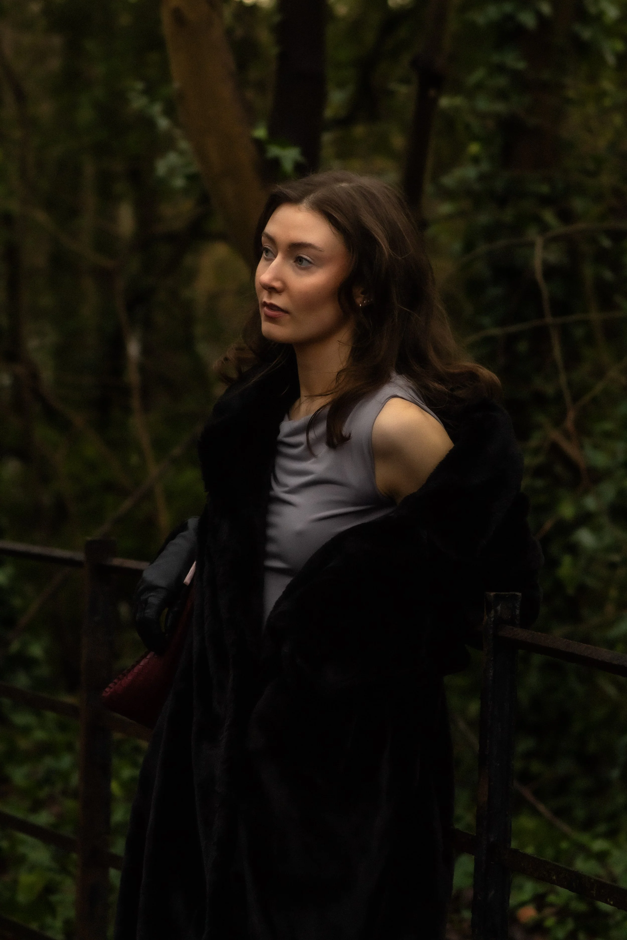 Young woman with wavy brown hair wearing a gray sleeveless top and a black coat, standing outdoors by a rusted metal railing in a wooded area.