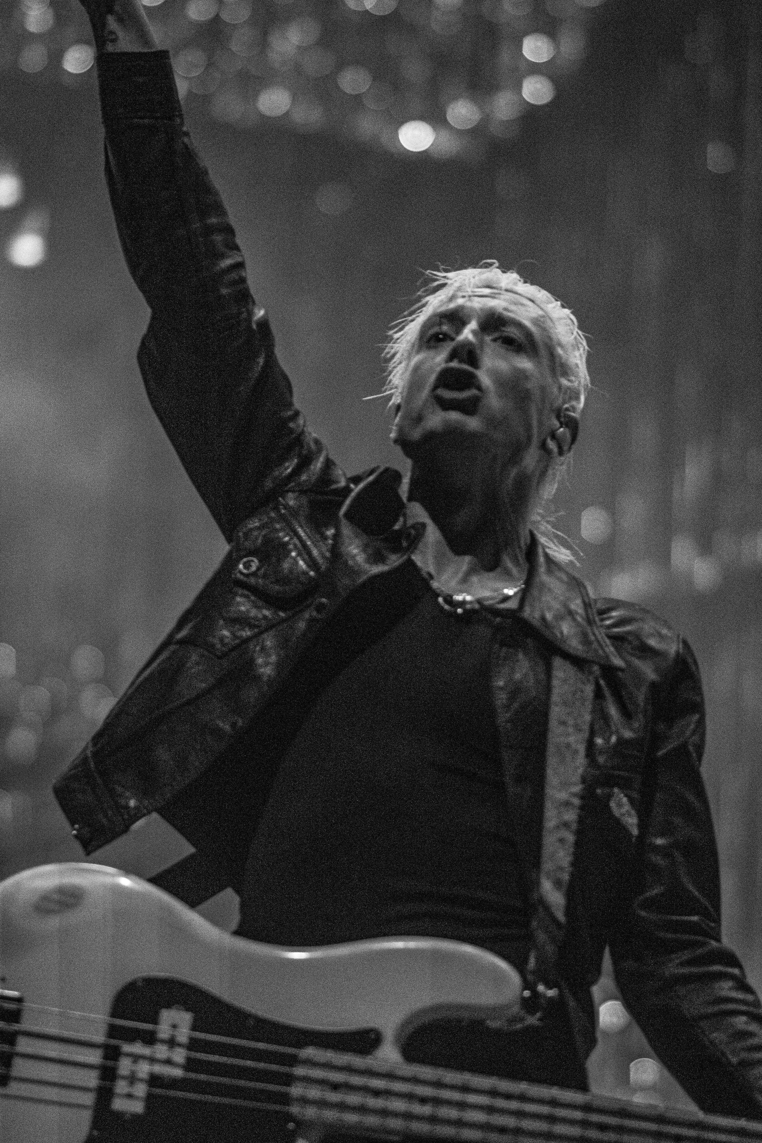 A black and white photo of a man performing on stage, holding a bass guitar, with his mouth open and finger pointing upward, wearing a leather jacket and necklace.