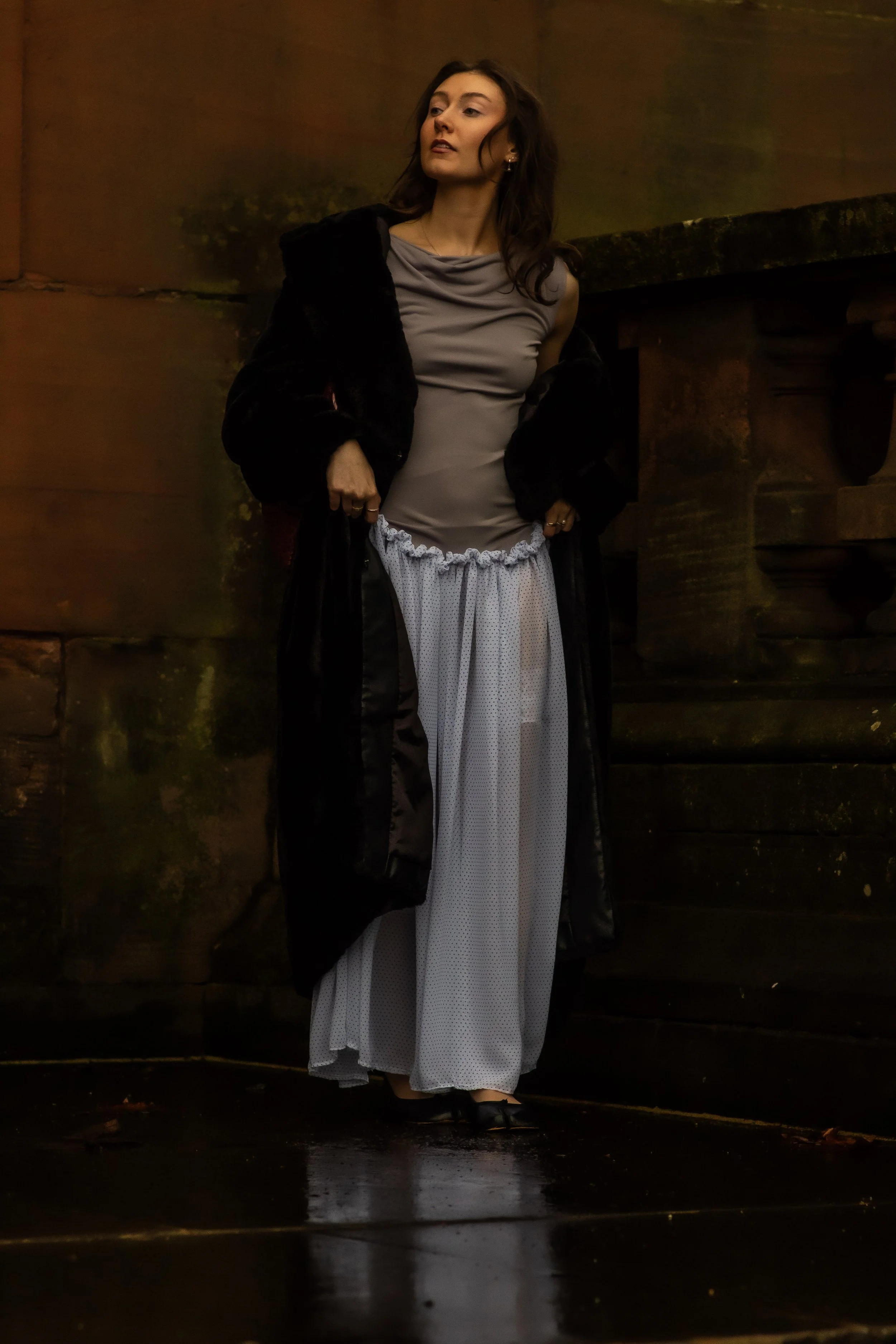 A woman stands against a stone wall, wearing a sheer white pant skirt, gray top, and a black coat draped over her shoulders, with a confident expression.