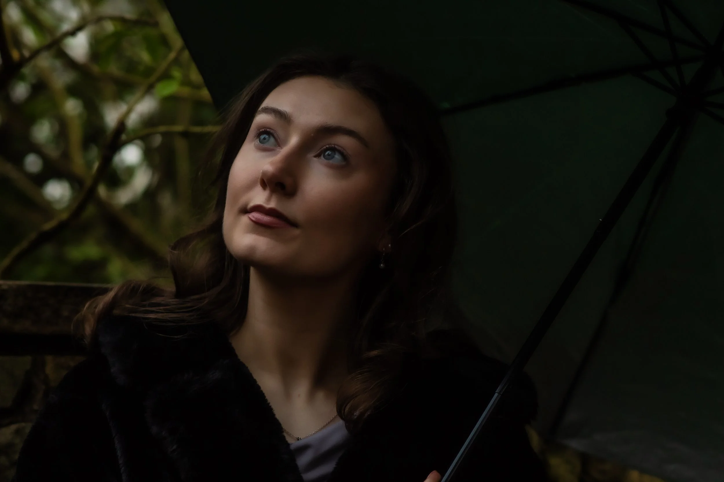 A woman with blue eyes and brown hair holding a dark green umbrella outdoors in a forest or garden setting.