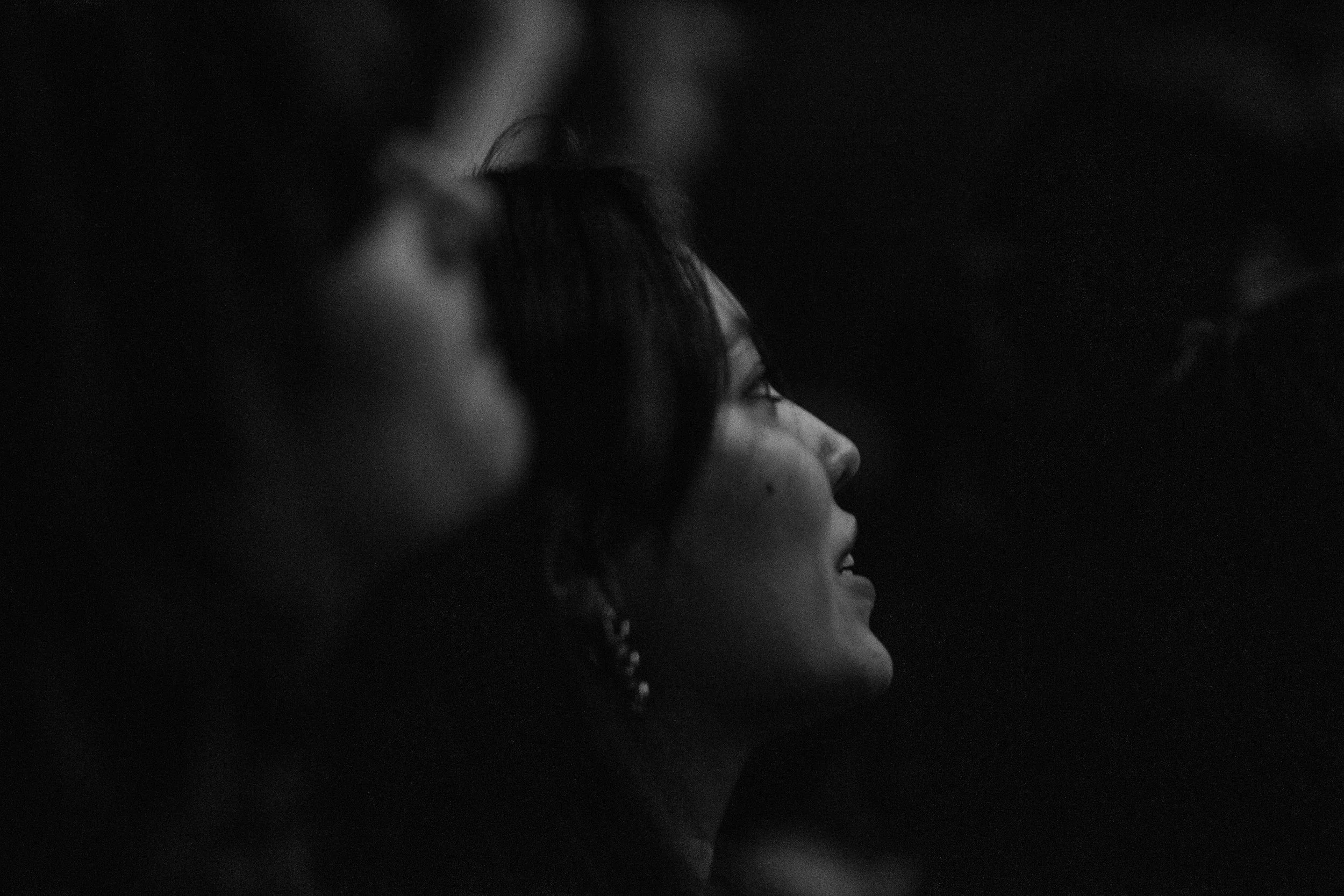 Side view of a smiling woman with dark hair and earrings, looking upward in a low-light setting.