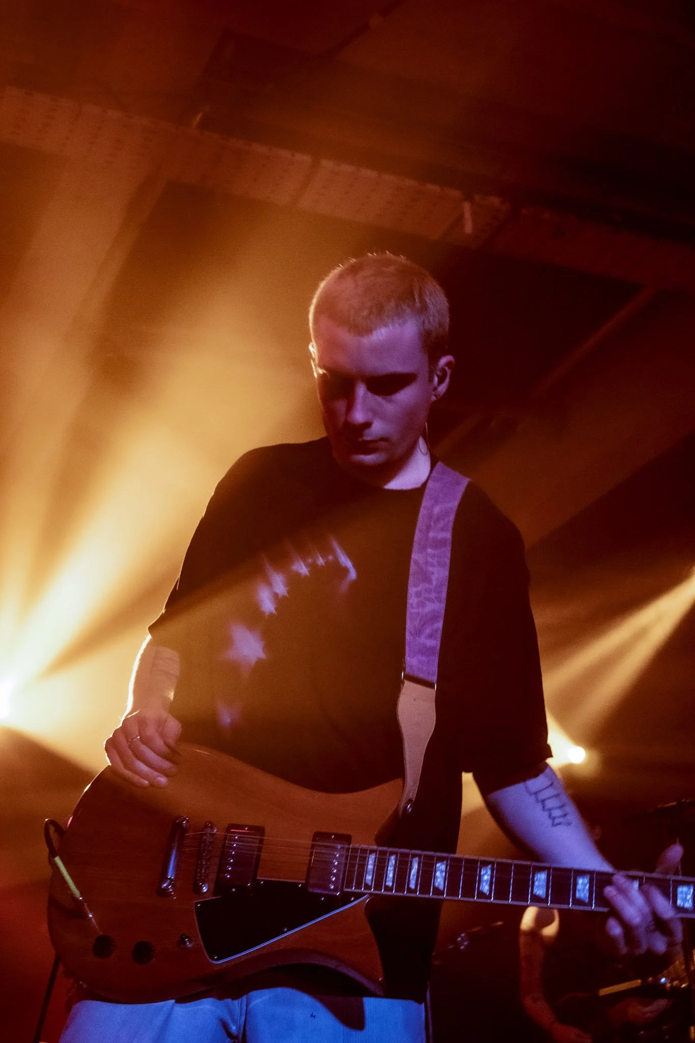 A young man playing an electric guitar on stage with orange and yellow lights shining behind him.