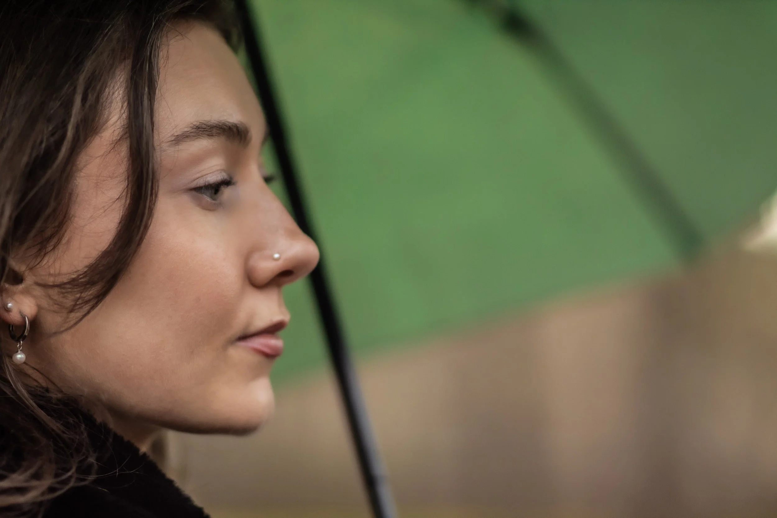 Profile of a young woman with dark brown hair, wearing earrings and a nose ring, holding a black umbrella outdoors with a green blurred background.