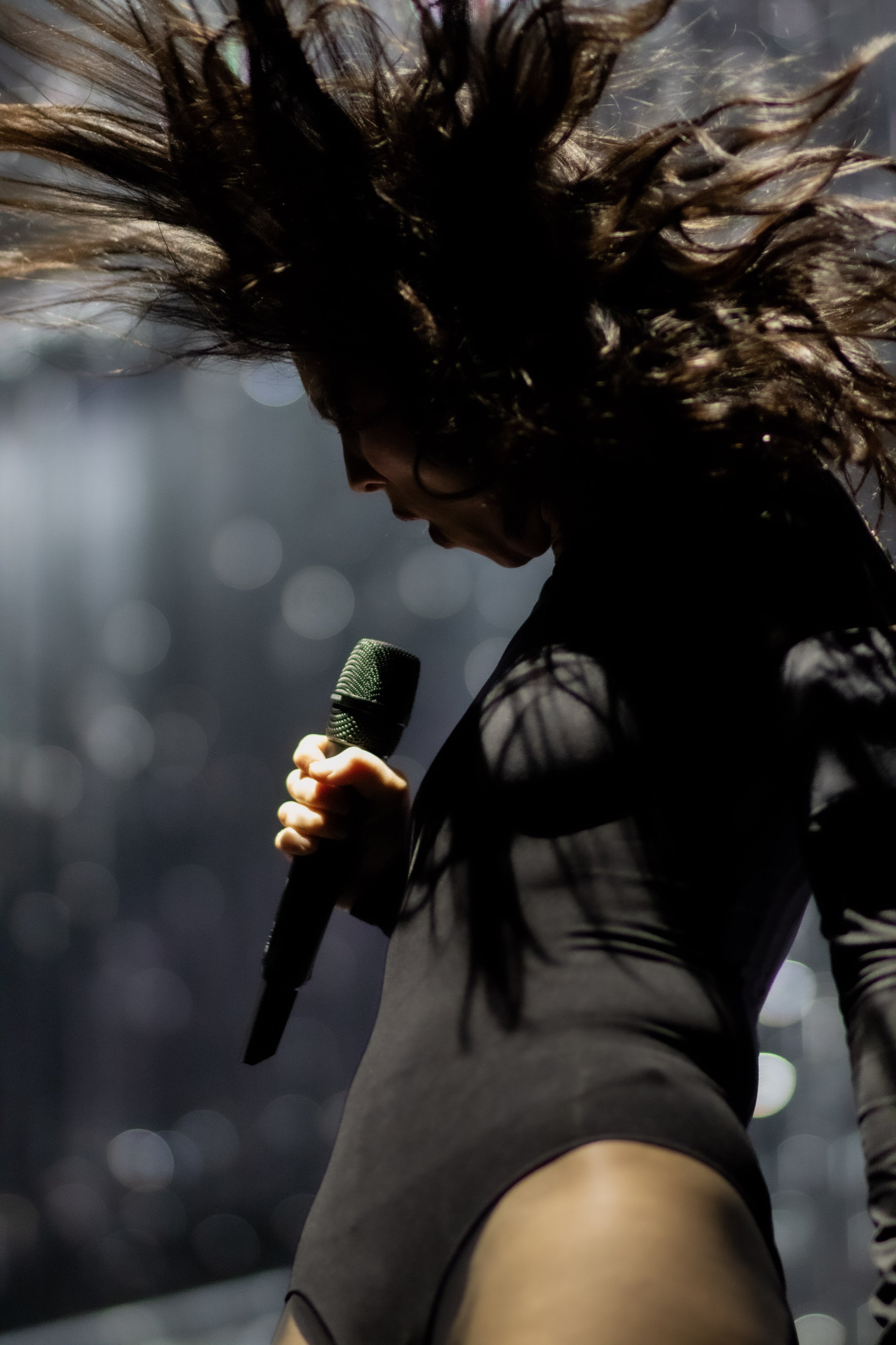 A female performer on stage, holding a microphone, with her eyes closed and hair flying around her face, wearing a dark outfit.