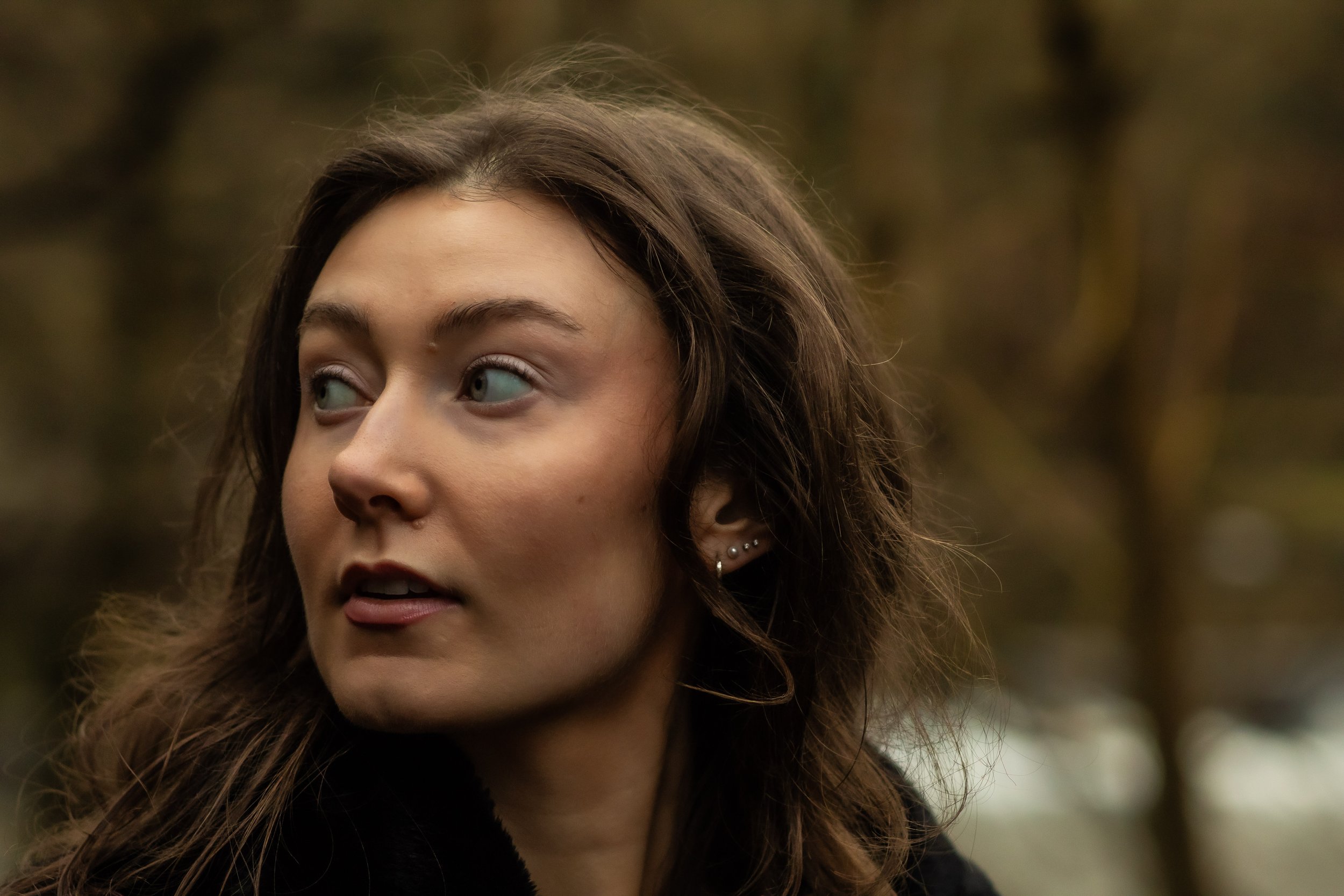 Close-up of a young woman with long, wavy brown hair and multiple ear piercings, looking to the side outdoors with blurred autumn trees in the background.
