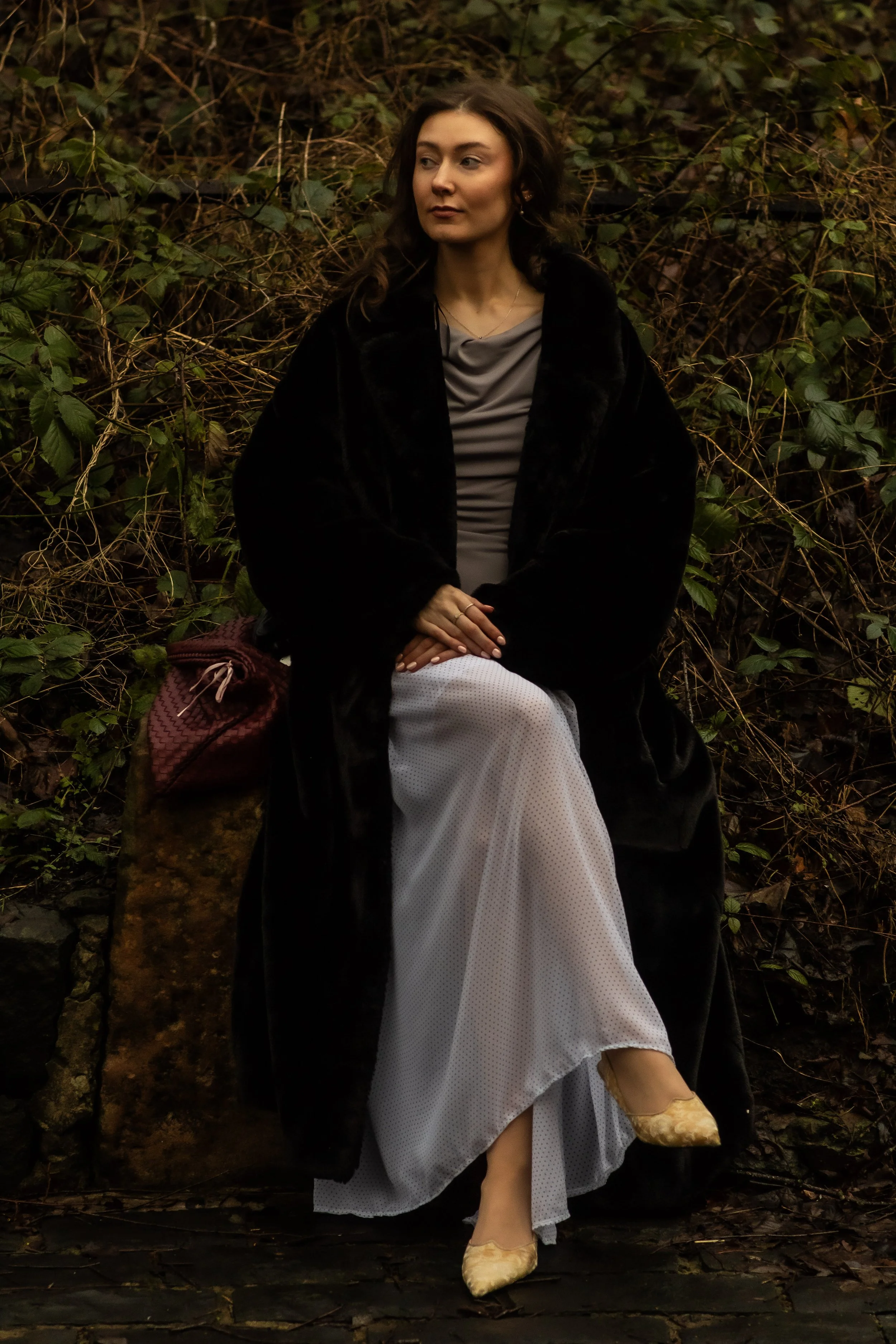 A woman sitting outdoors on a stone ledge, with a background of dense greenery. She has long, wavy brown hair and is wearing a gray top, a long white skirt, and a black coat. She has a maroon bag beside her on the stone, and she is wearing beige shoe