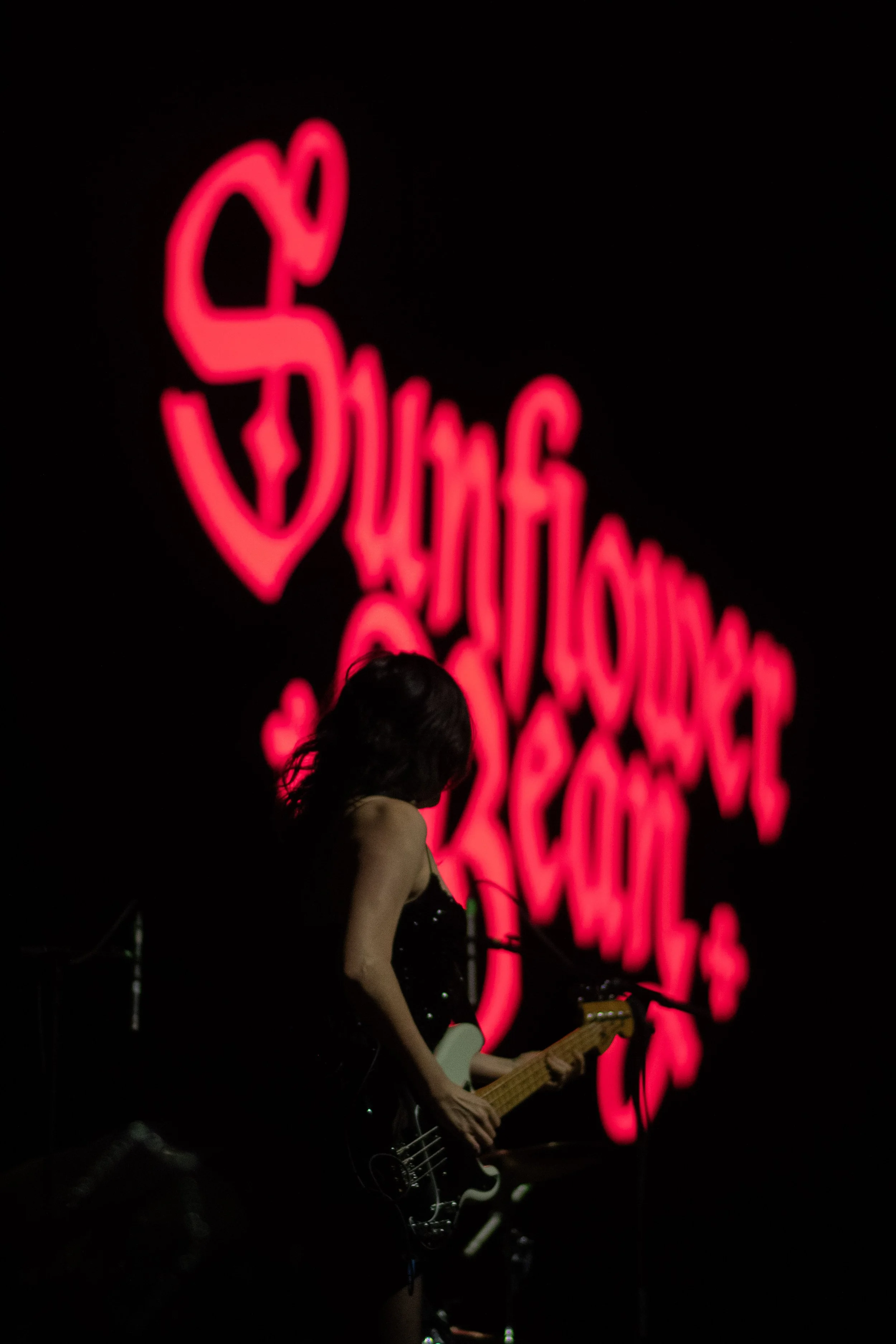 A person playing an electric guitar on stage with a black background and a large, glowing neon pink sign in the background.