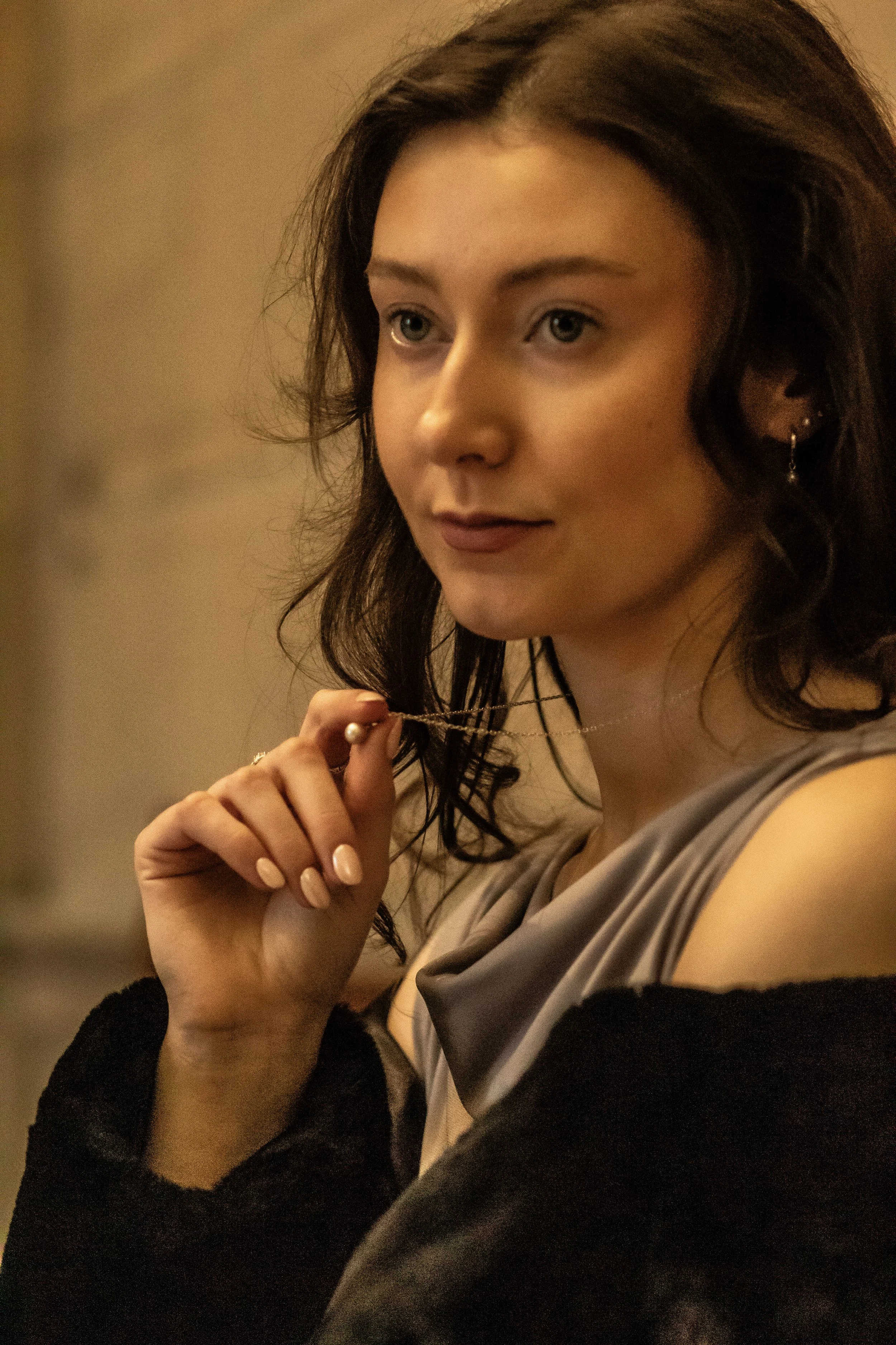 A woman with wavy dark hair, wearing earrings and a gray top with a black garment, is holding a necklace with a pearl pendant near her face, looking thoughtfully to the side.