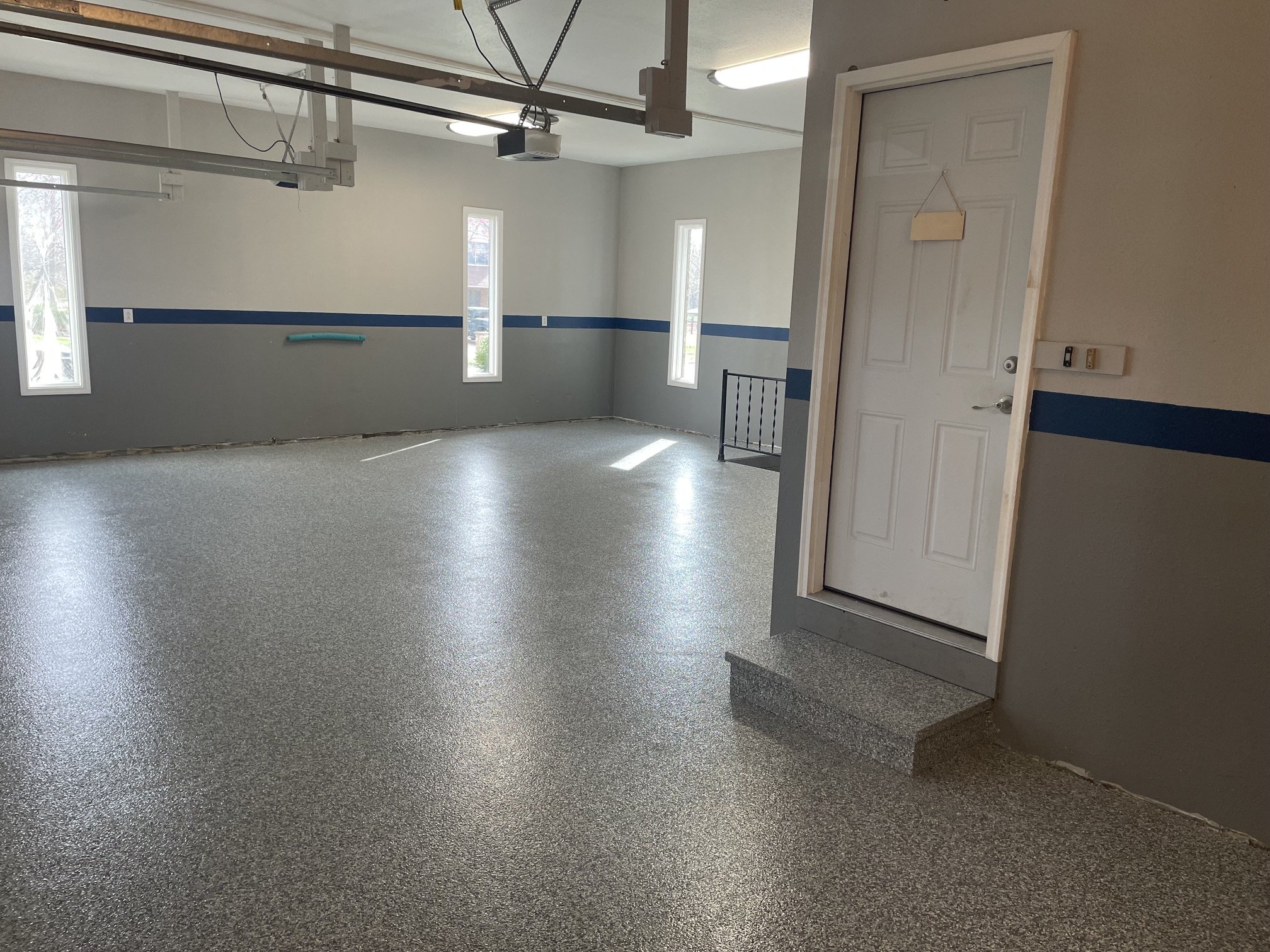 Clean garage with gray speckled epoxy flooring, white walls, and multiple narrow windows letting in natural light. There is a white door with a small hanging sign, and some garage equipment and wiring are visible at the ceiling.