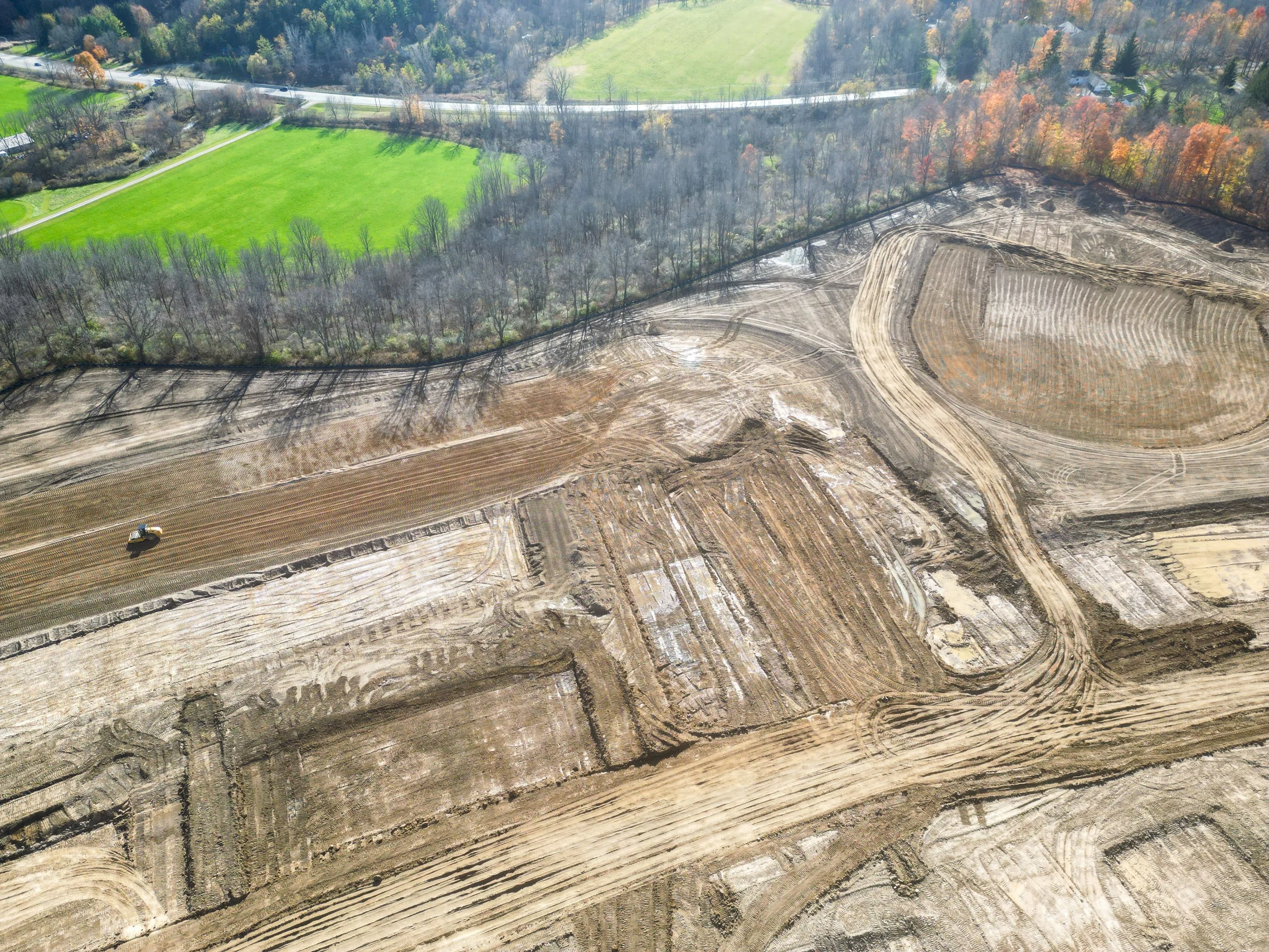 Aerial view of a construction site with cleared land, dirt roads, and a bulldozer, adjacent to a forested area with trees and green fields.