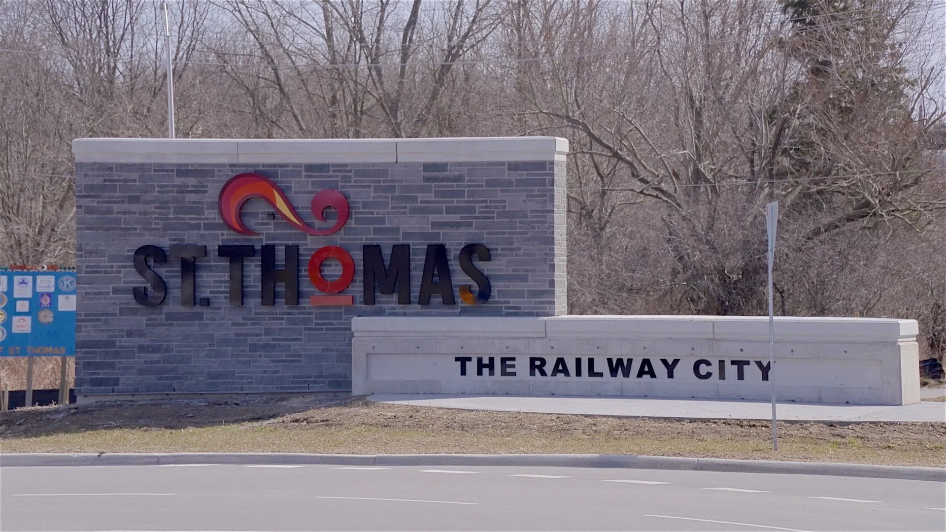 Sign for St. Thomas, The Railway City, with a decorative logo and a stone wall, situated beside a road with leafless trees in the background.