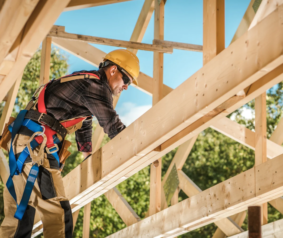 A construction worker wearing a yellow hard hat, safety glasses, and a harness is working on a wooden building frame.
