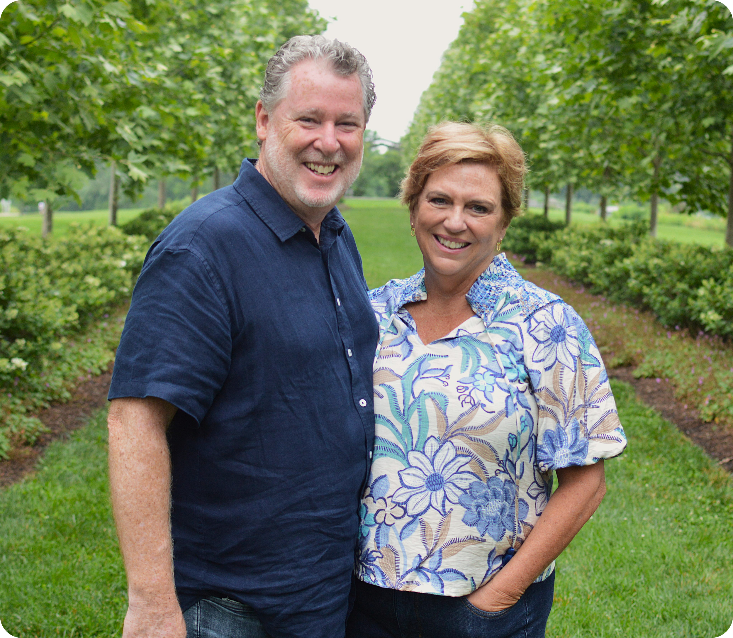 A smiling middle-aged man and woman standing outdoors in a lush green park with trees and bushes, posing for the photo.
