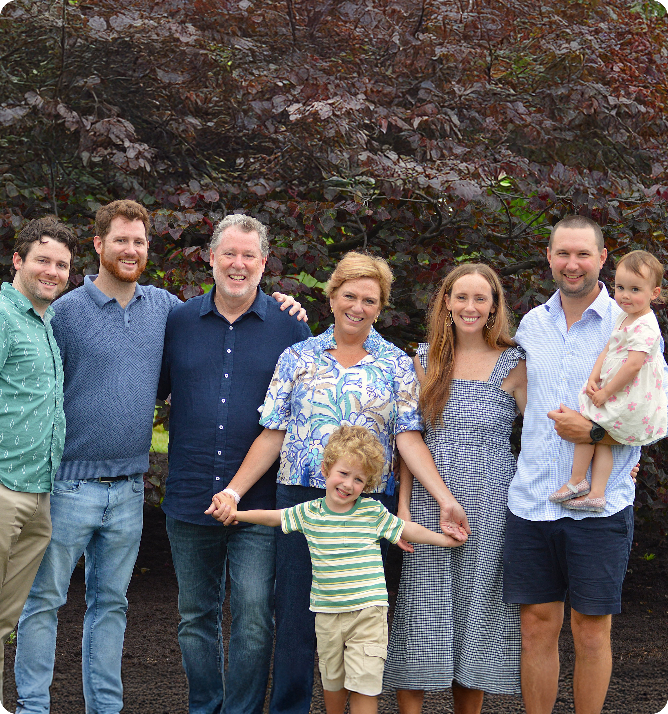 Family portrait outdoors in front of a dark leafy tree, including seven adults and two children, all smiling and holding hands.