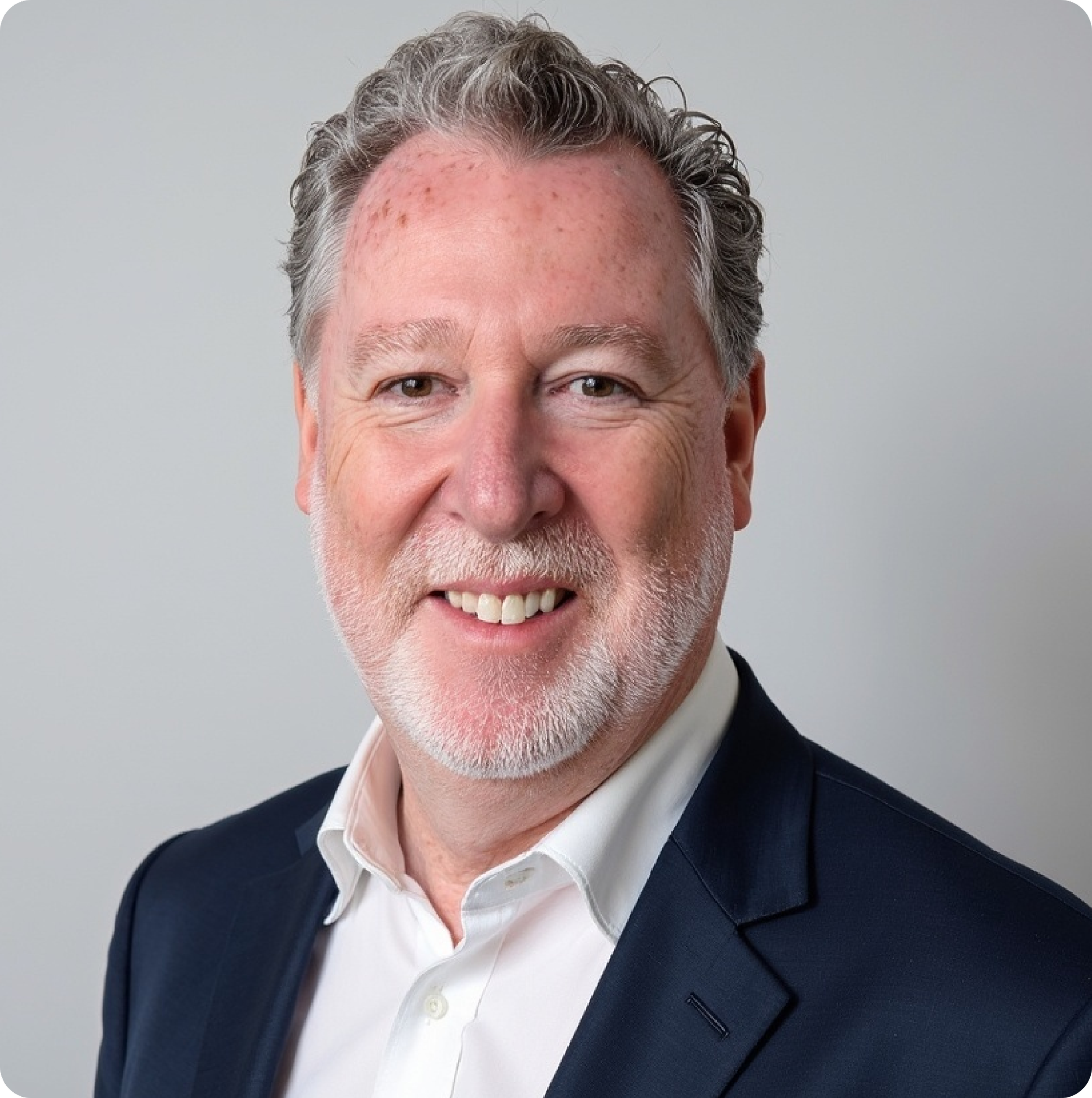 Headshot of a middle-aged man with gray hair and a beard, wearing a navy blazer and white shirt, smiling against a plain gray background.