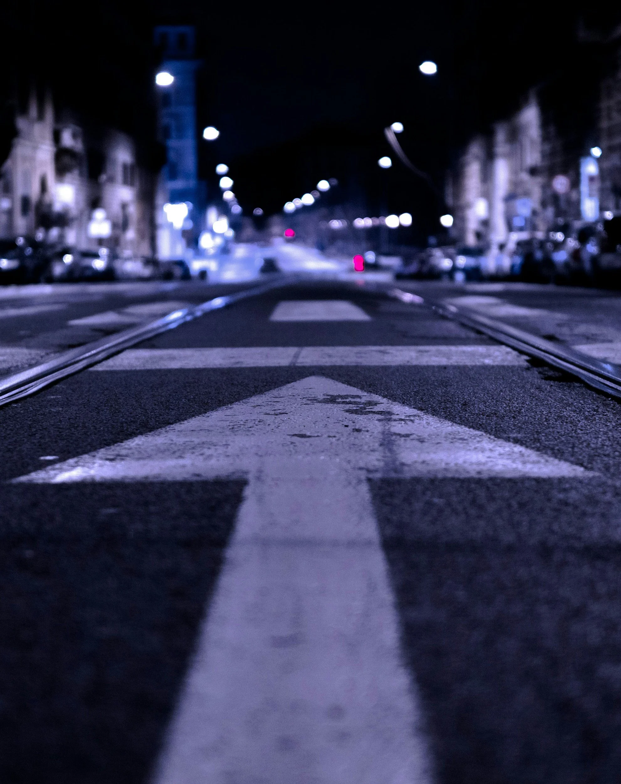 Nighttime street scene with a painted white arrow on the road pointing forward, tram tracks on both sides, illuminated buildings, and streetlights.