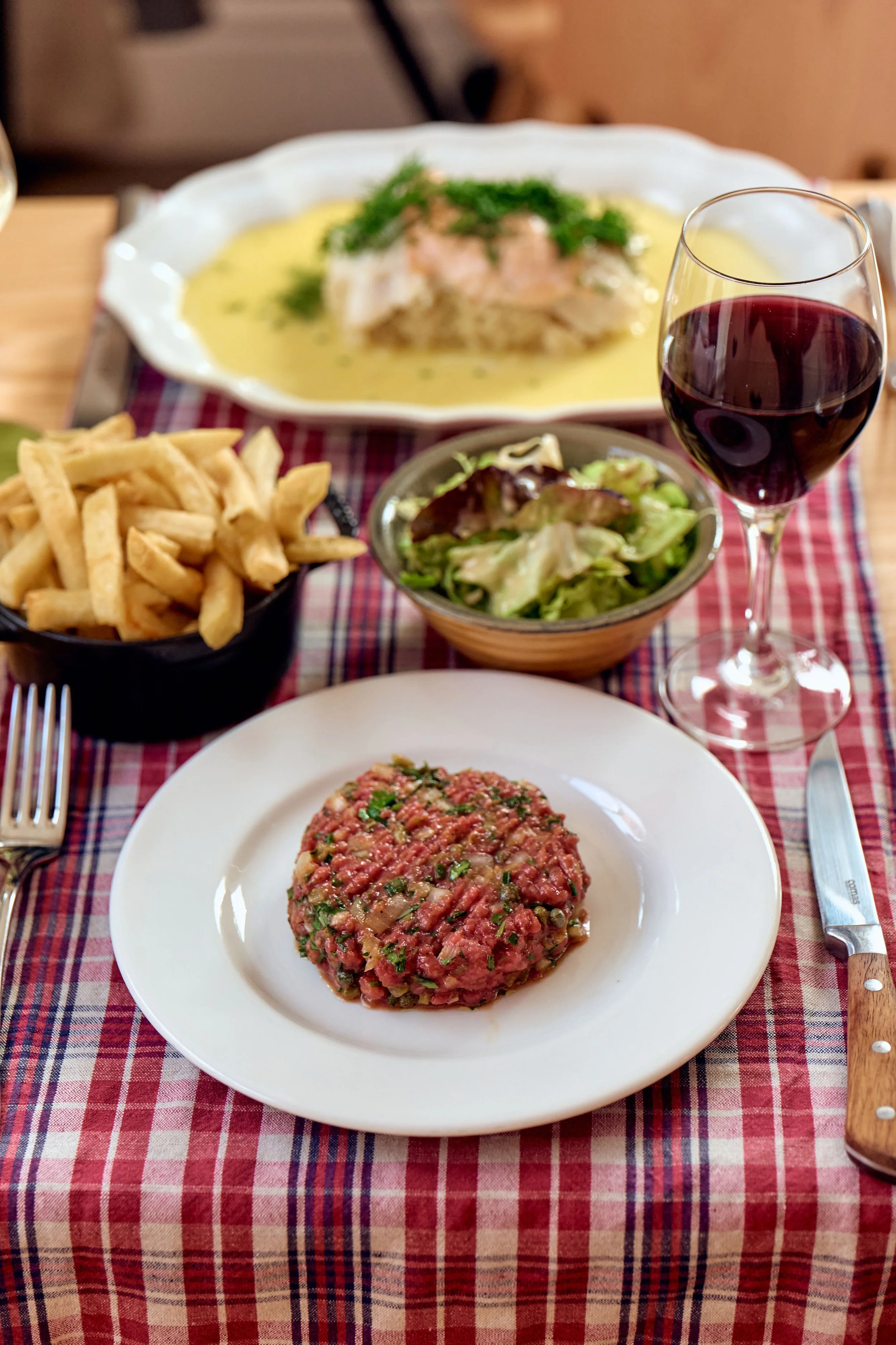 A table set with French fries, a bowl of salad, a glass of red wine, and a plate of steak tartare, with a baked fish dish in the background on a red checkered tablecloth.