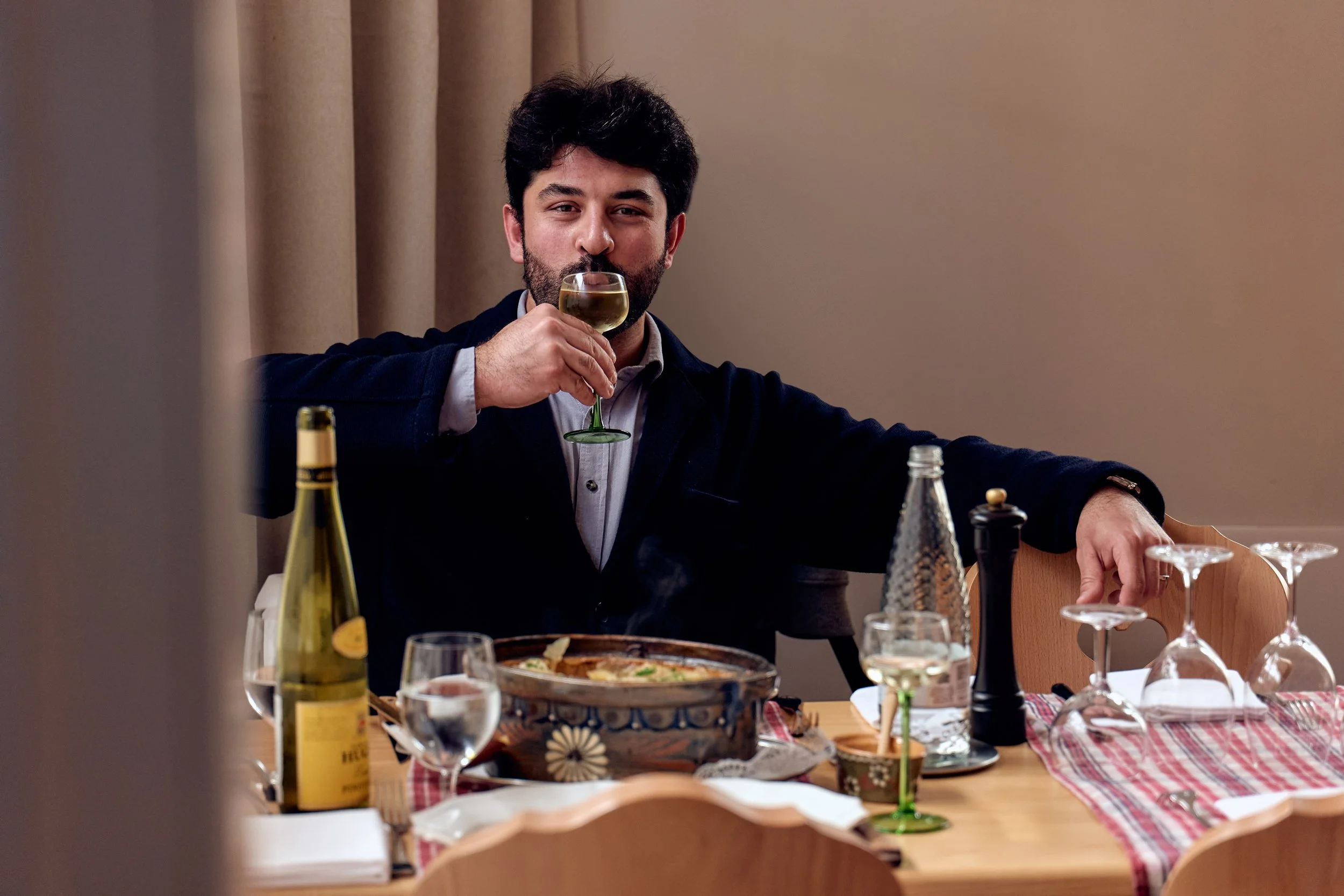 Man sitting at a dining table, drinking white wine, with a covered dish and wine bottles on the table.