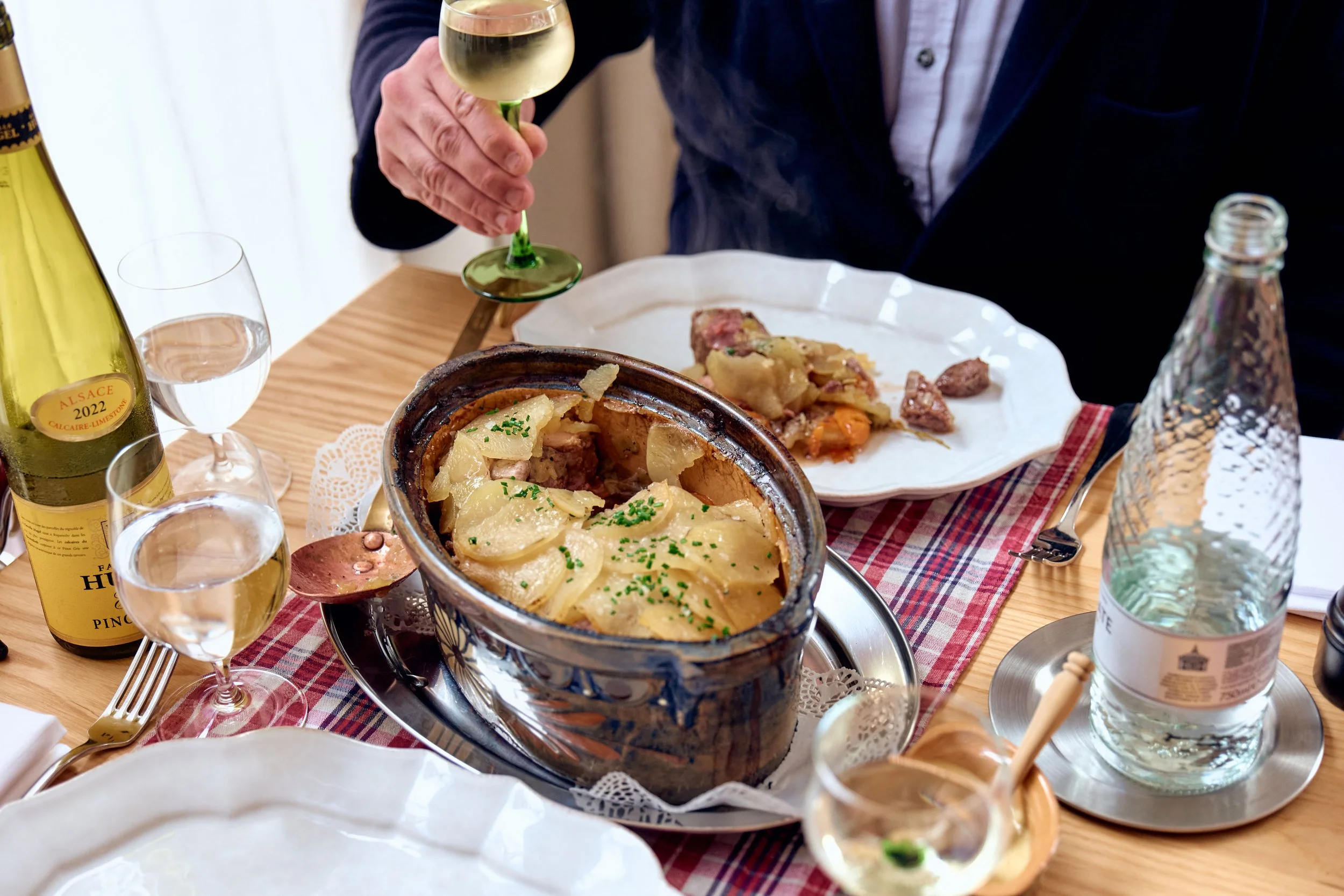 A person holding a glass of white wine at a dining table set for a meal, with a pot of potato gratin, plates of food, bottles of water and wine, and utensils on a red checkered tablecloth.