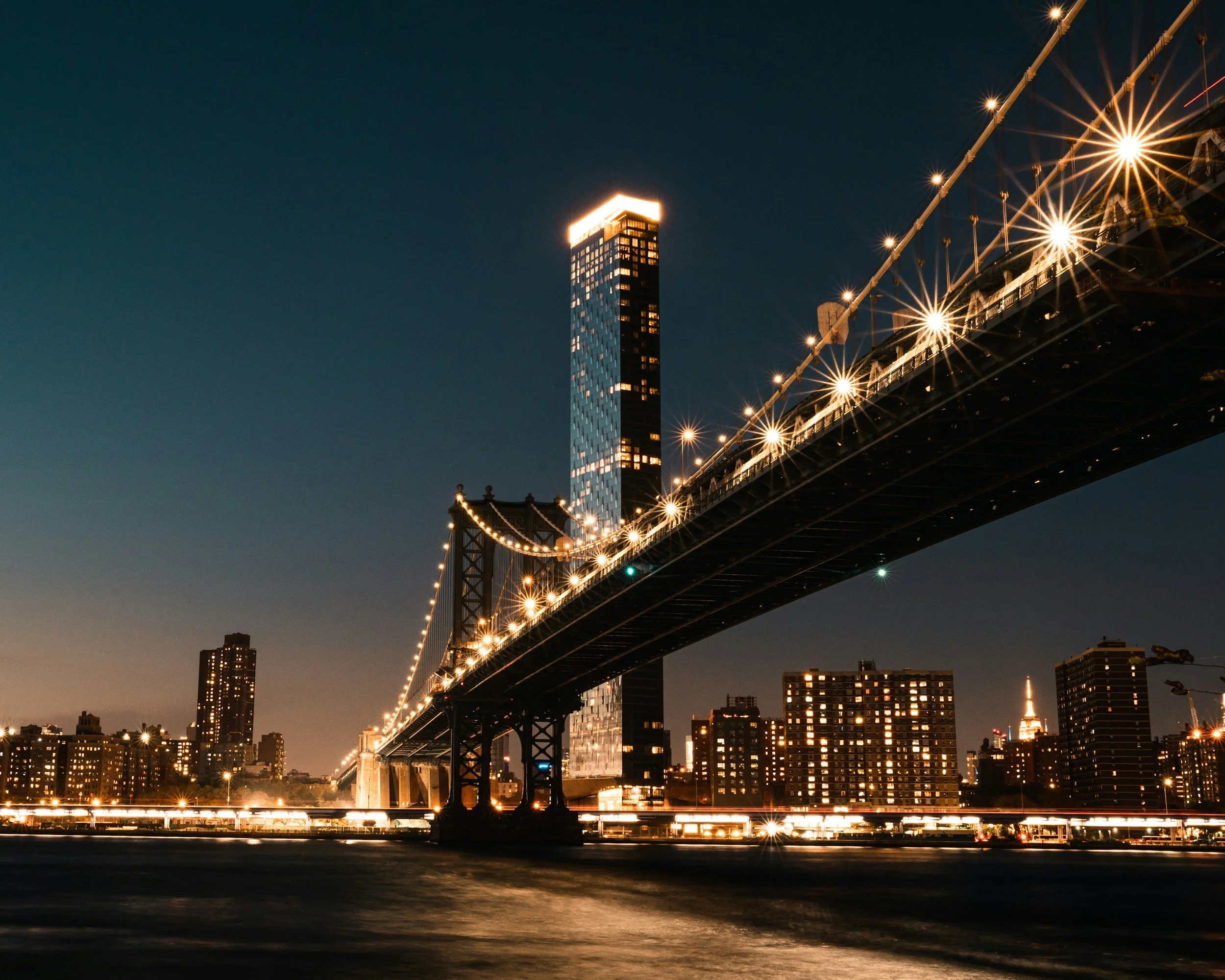 Night view of a city skyline with a lit-up suspension bridge in the foreground, and a modern skyscraper with illuminated top in the background, with the Eiffel Tower visible in the distance.