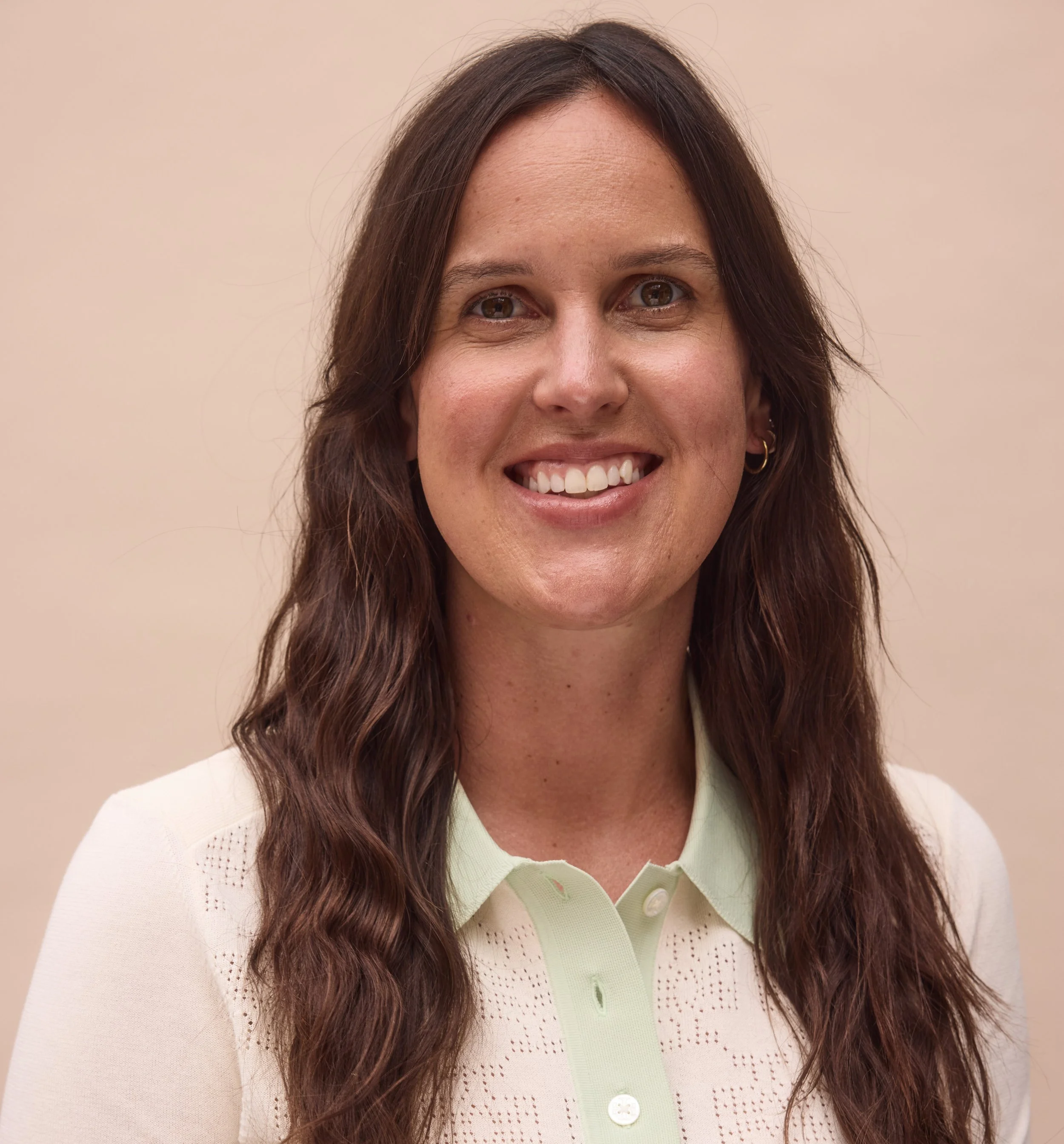 A woman with long wavy brown hair, smiling, wearing a light green collared shirt and small hoop earrings, standing against a plain beige background.
