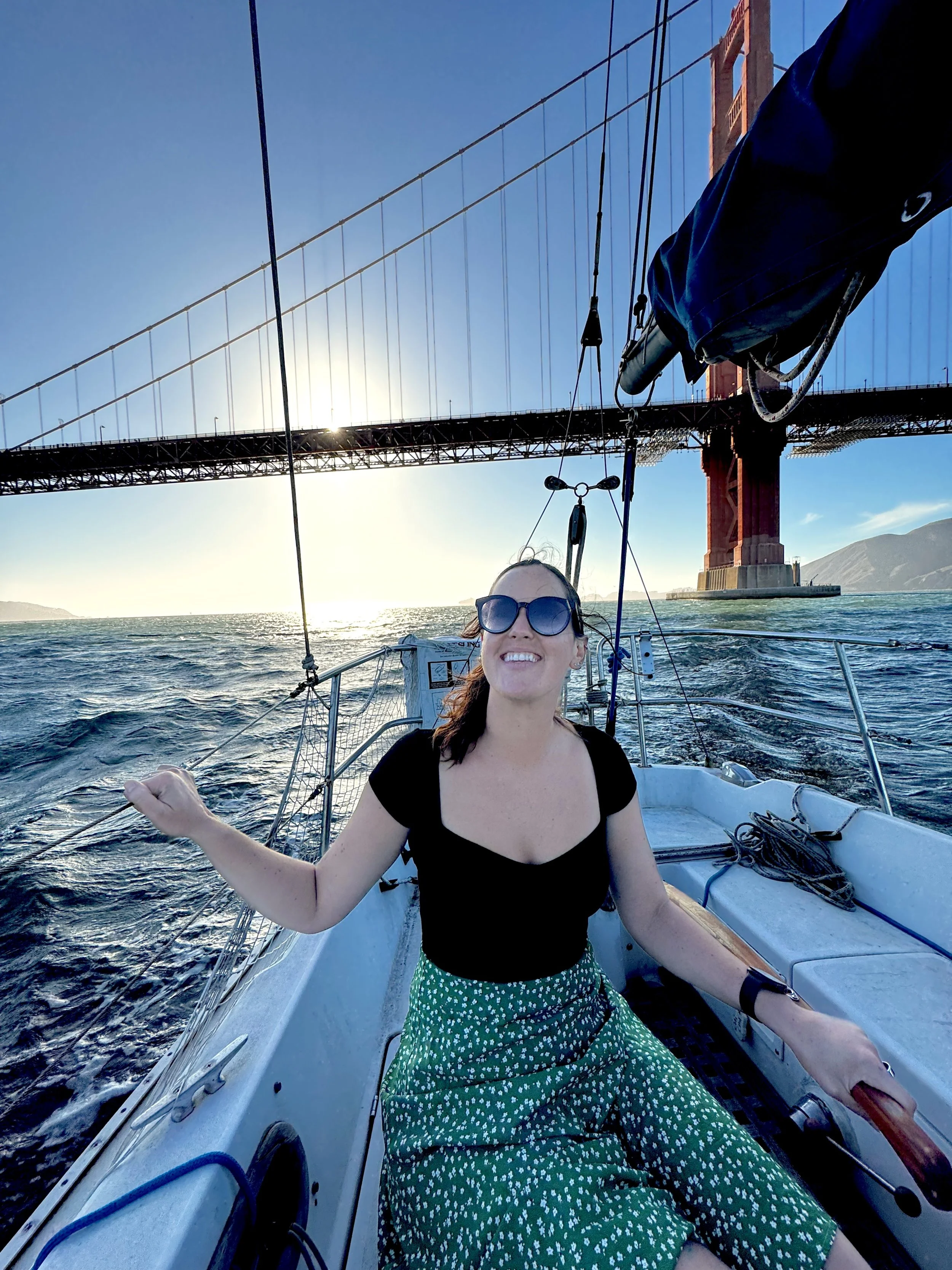 Woman in sunglasses and a black top smiling on a sailboat with the Golden Gate Bridge in the background.