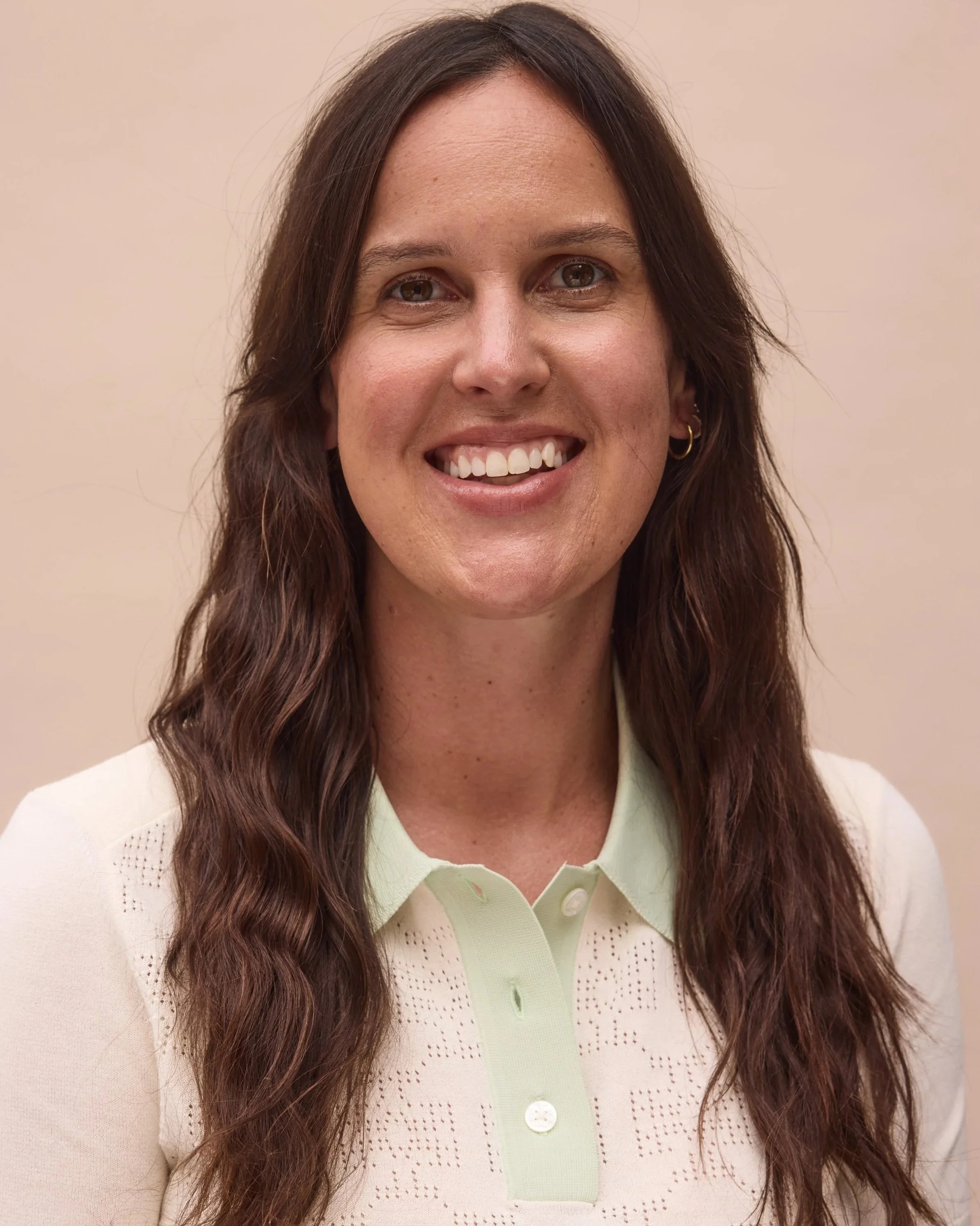A woman with long wavy brown hair, wearing a light-colored collared shirt, smiling and standing against a plain beige background.