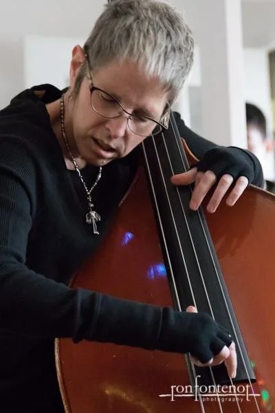 Person with short blond hair and glasses playing a double bass, wearing a black shirt and gloves, in an indoor setting.
