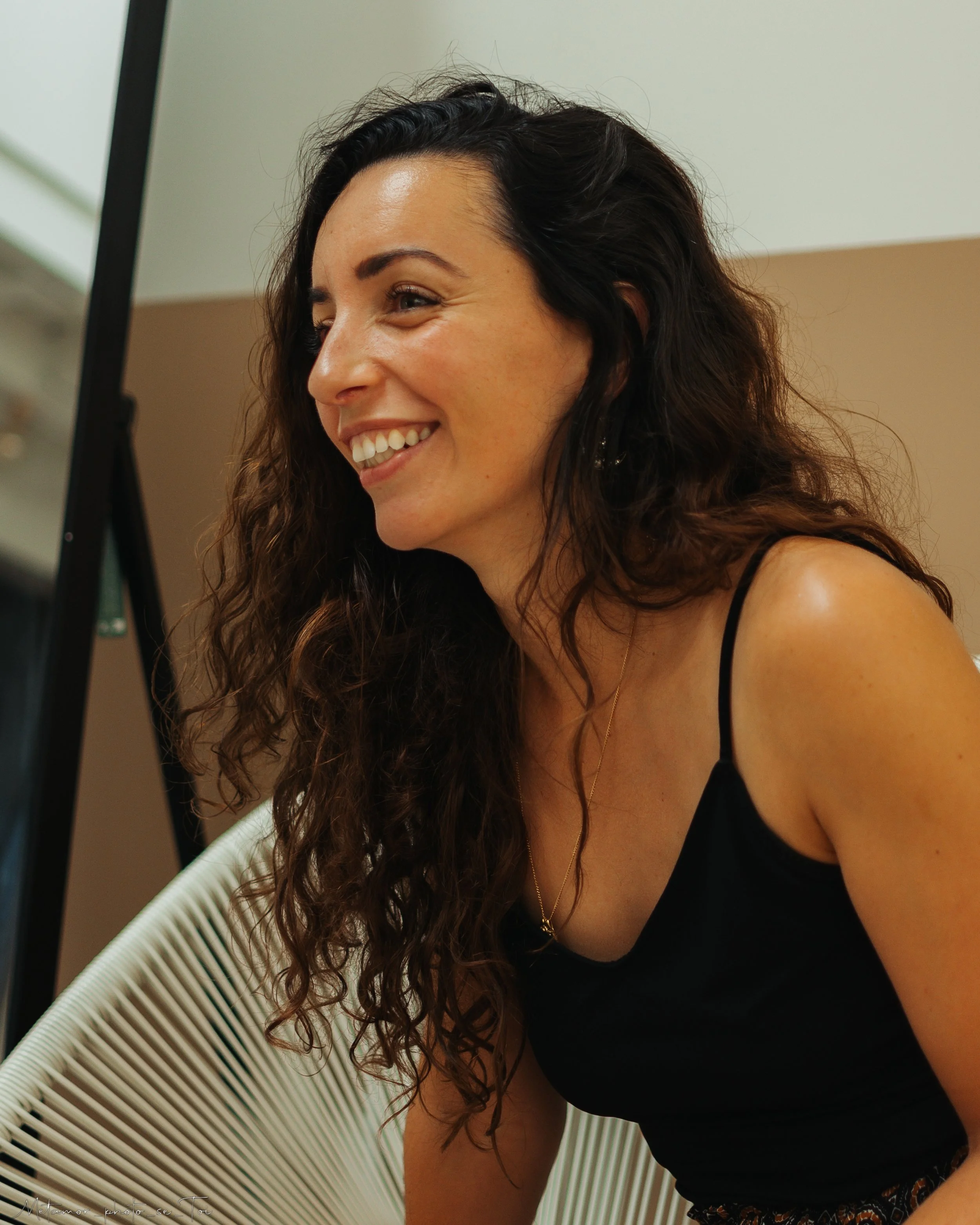 Une femme souriante avec des cheveux bouclés bruns assise sur une chaise