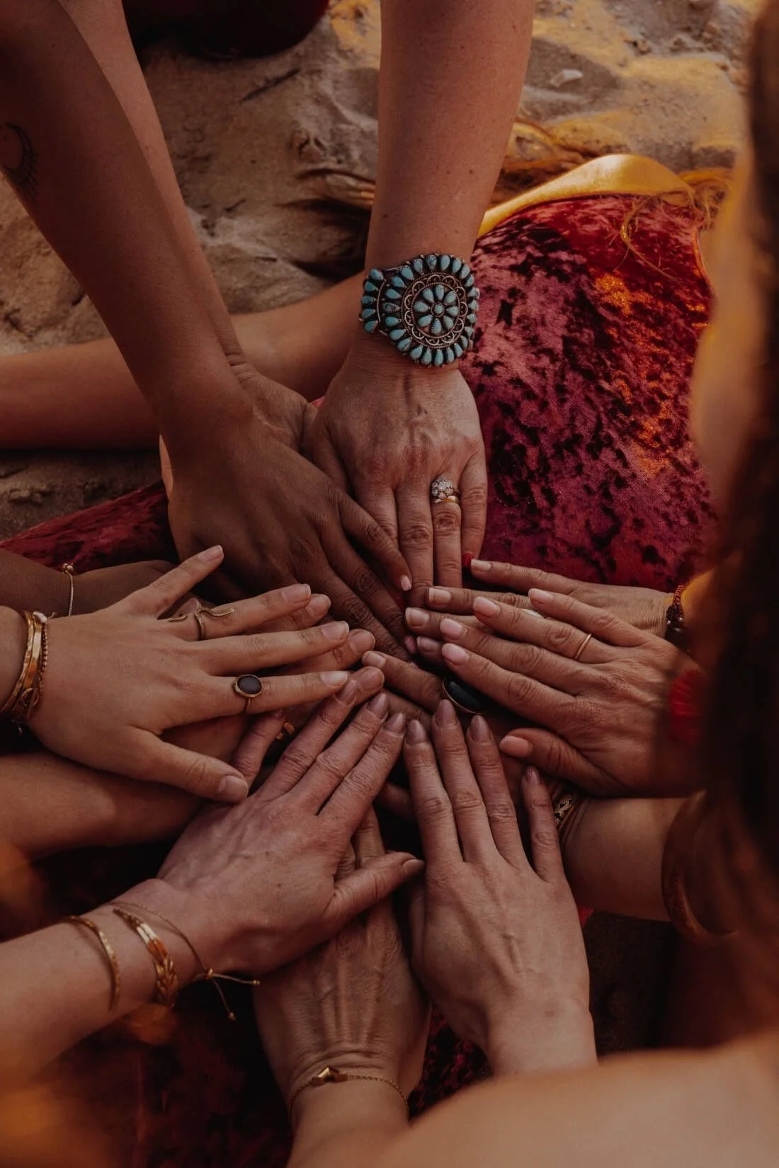 Groupe de mains réunies en cercle, certaines portent des bijoux, sur un fond de tissu orange et rouge.