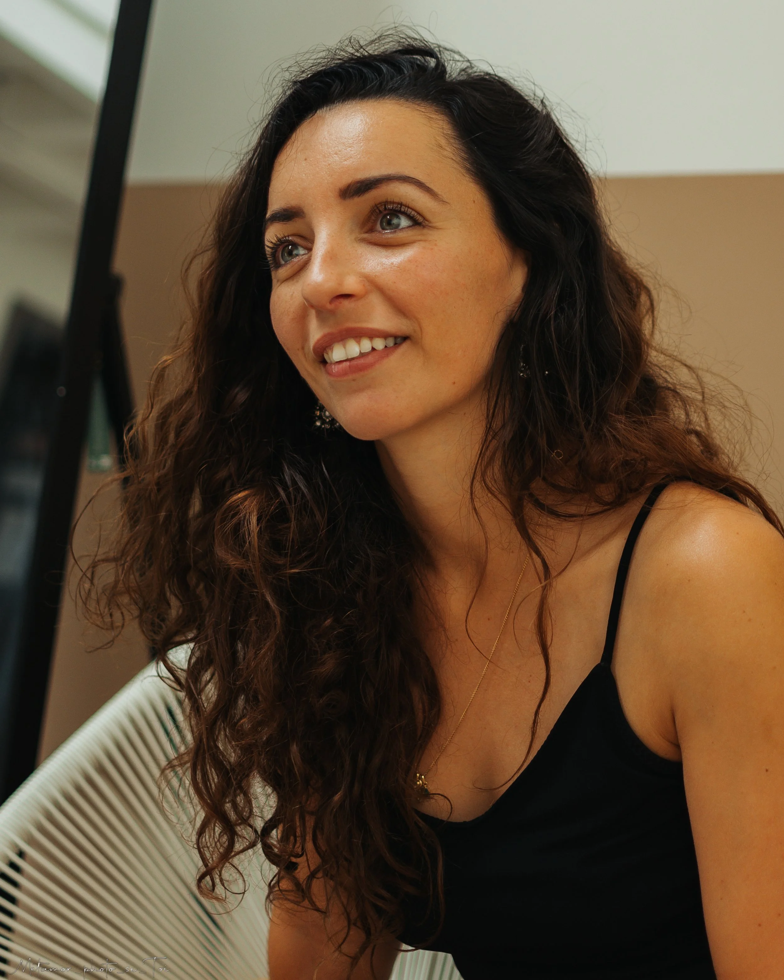 Jeune femme aux cheveux bouclés assise, souriant, portant un débardeur noir, dans un environnement intérieur.