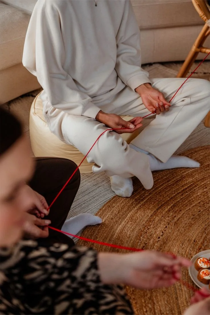 Deux personnes jouent à un jeu de ficelle, assises sur un tapis en intérieur.