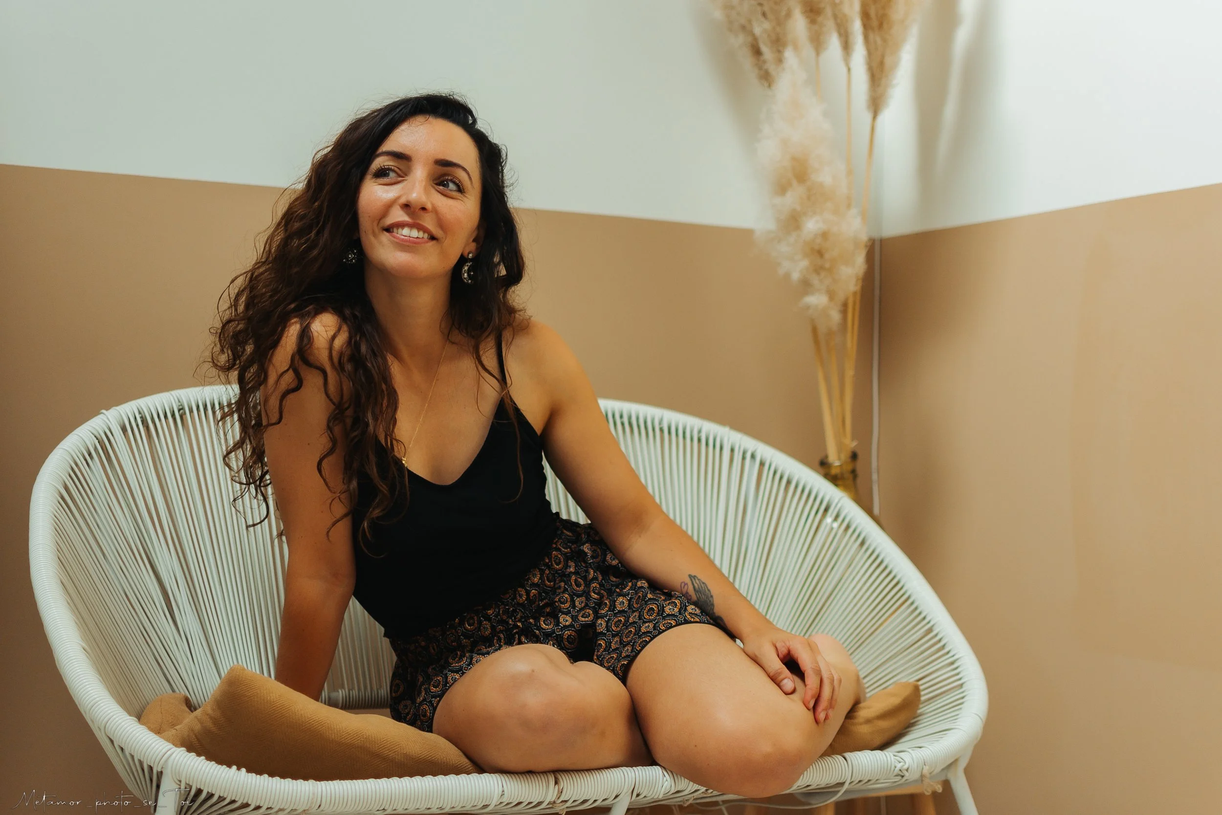 Une femme aux cheveux bouclés, souriante, assise dans un fauteuil en rotin blanc, portant un haut noir et un short à motifs colorés, dans une pièce décorée avec des plantes en plumes dans un vase en arrière-plan.