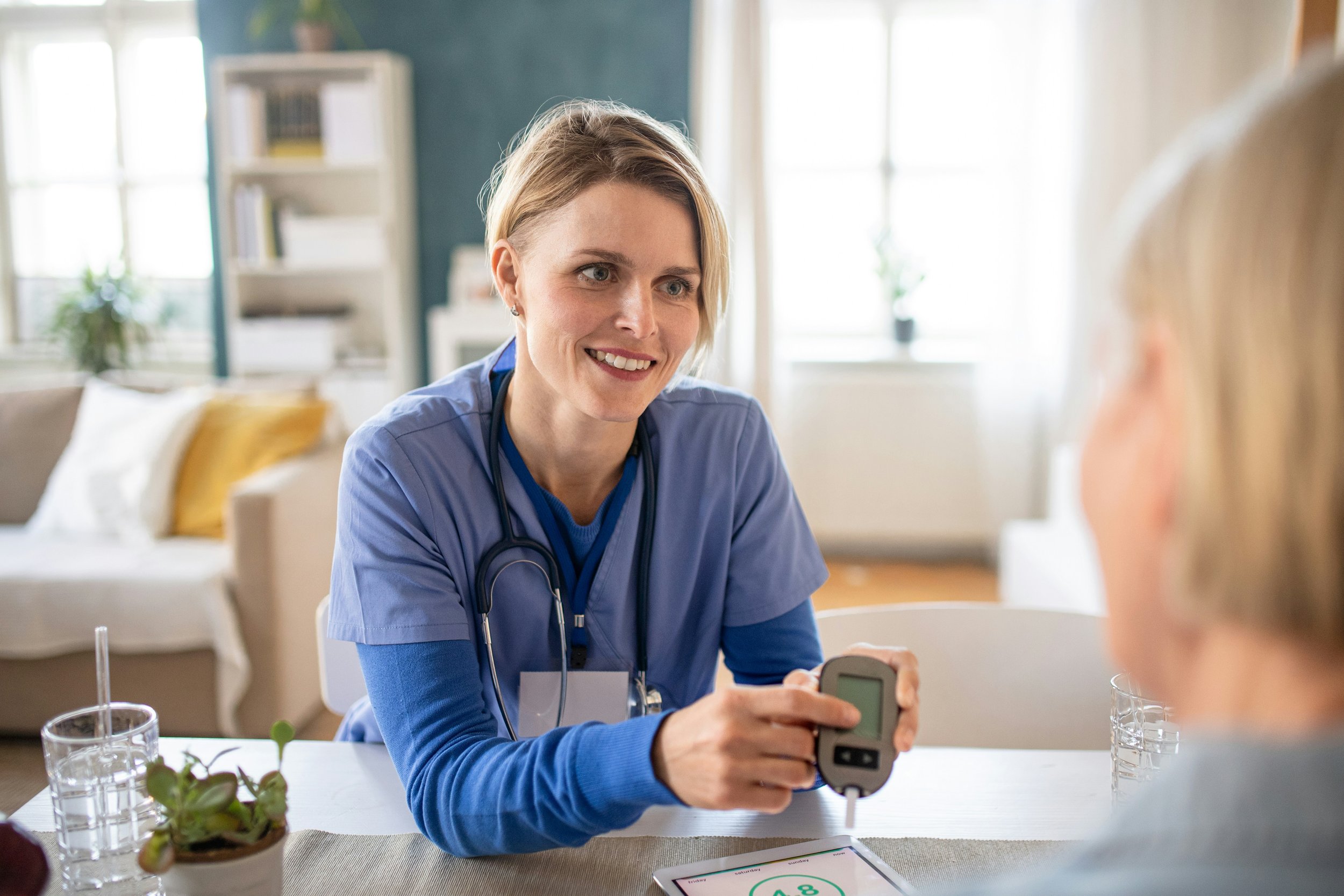 A woman wearing blue medical scrubs and a stethoscope around her neck, smiling, as she administers a glucose meter test to a patient in a well-lit room.