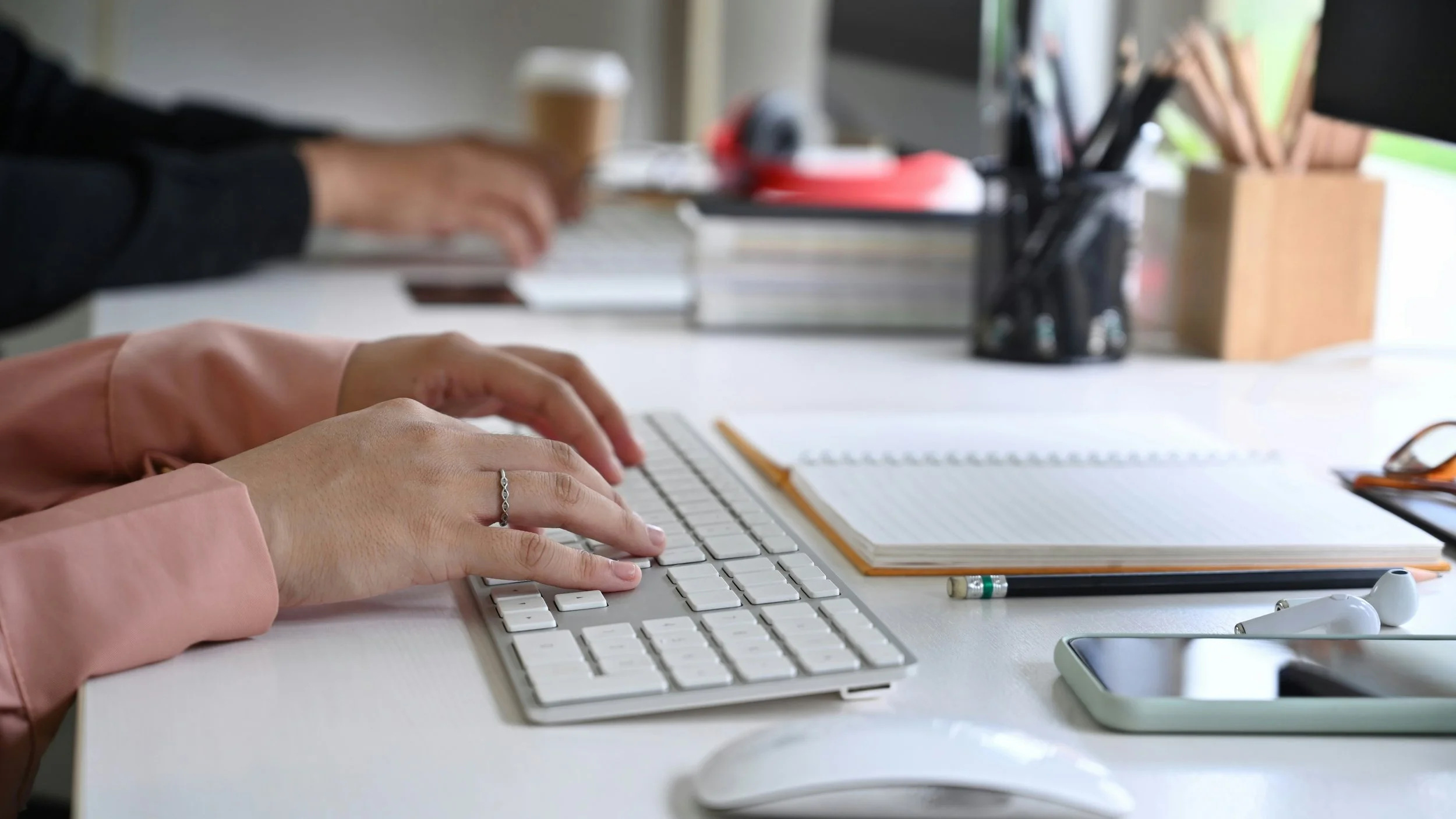 Person typing on a white keyboard with a pink long-sleeve shirt and a silver ring on their finger, on a white desk with notebooks, pens, and scattered office supplies.