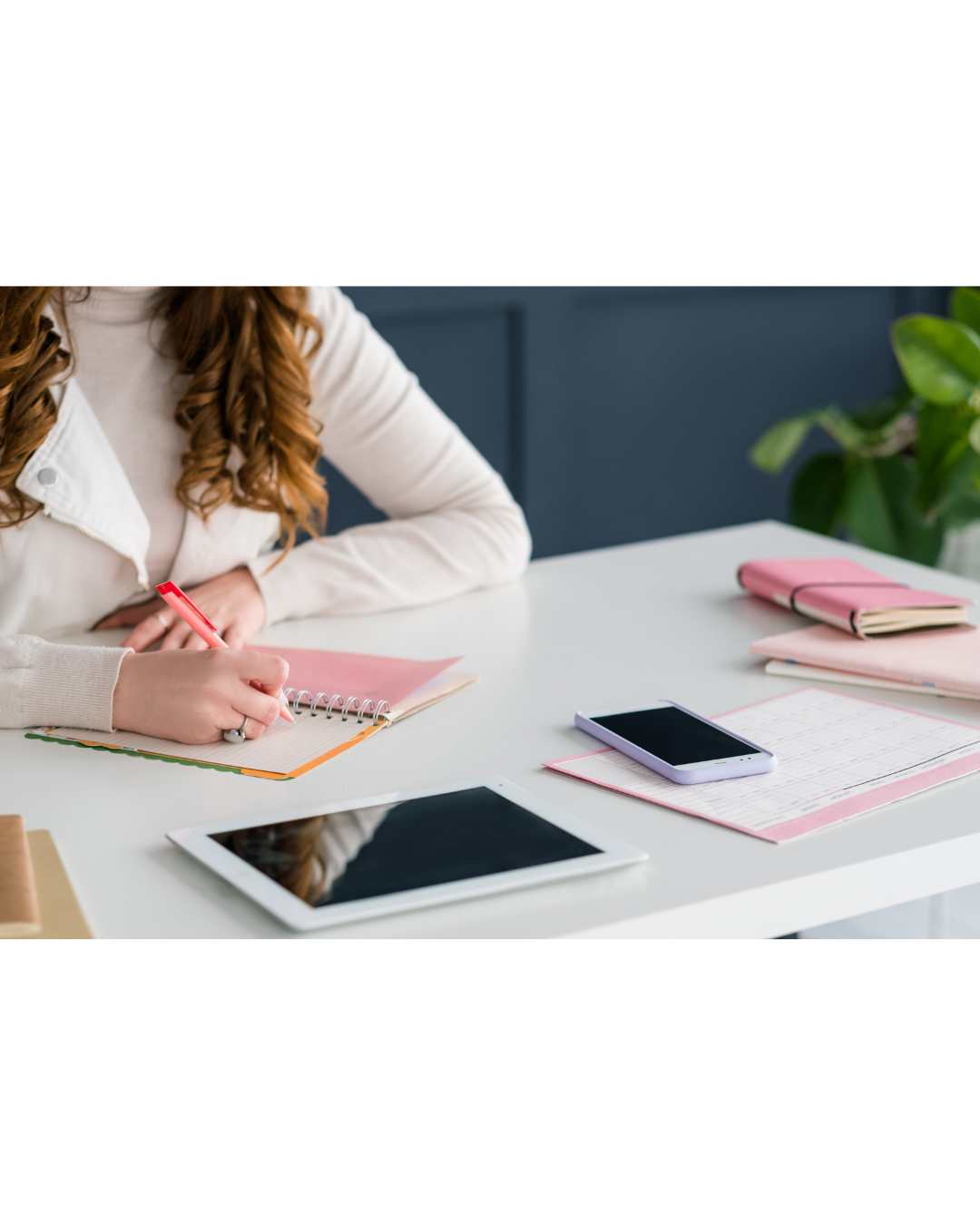 A person taking notes on a spiral notepad with a red pen on a white desk with electronic devices and pink notebooks.