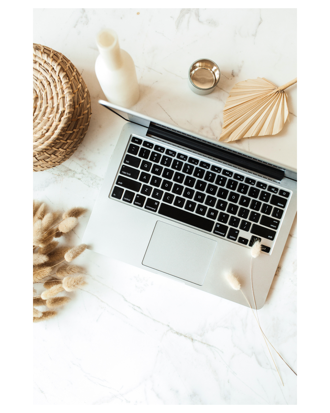 A flat lay of a workspace on a white marble surface, featuring a silver laptop with black keys, a woven basket, a white bottle, a small metallic container, an ivory paper fan, some dried flowers, and a few loose straw-like decorations.