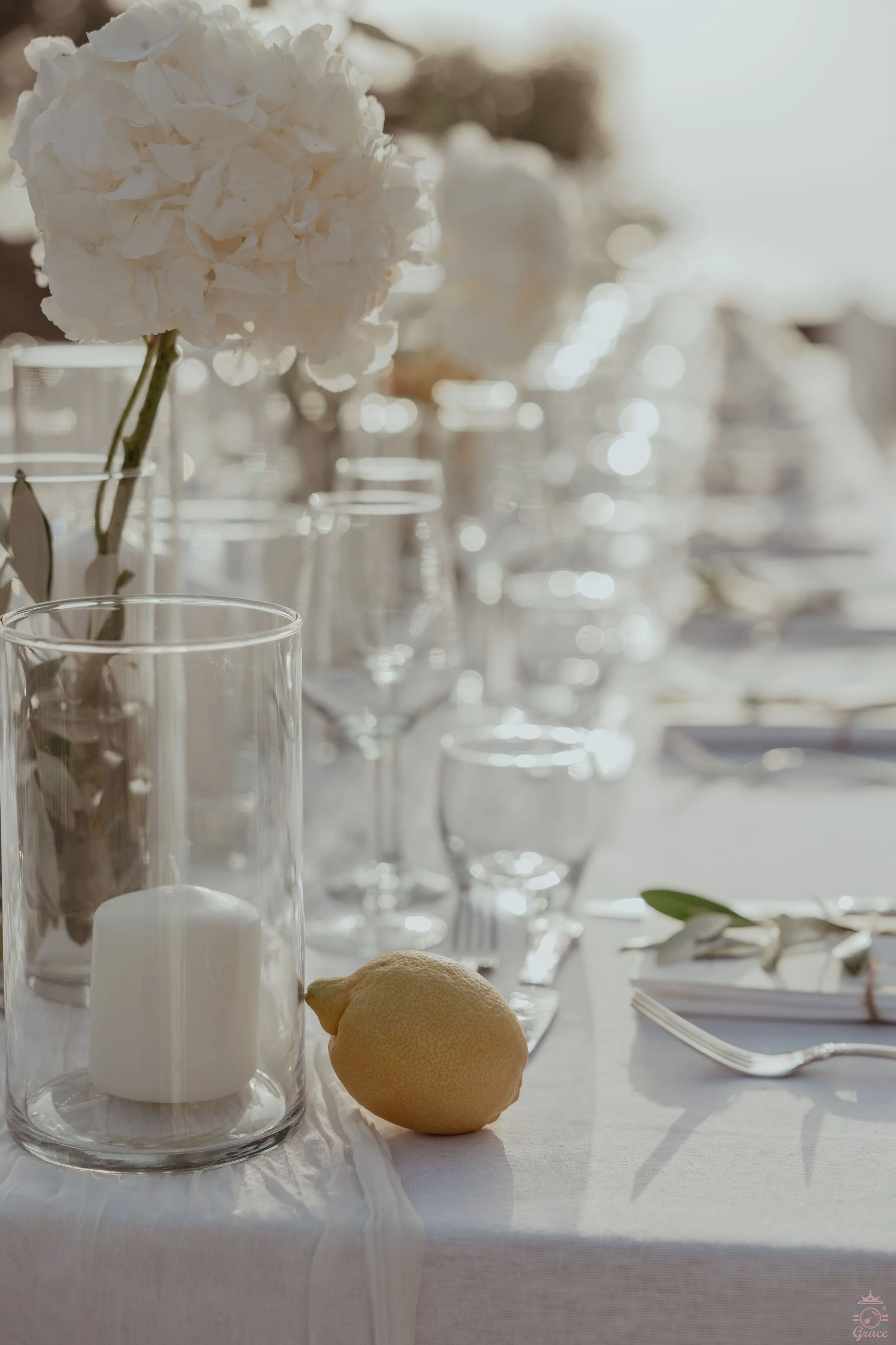 Table élégante décorée avec une fleur blanche, une bougie dans un vase en verre et un citron sur une nappe blanche, prêtes pour un repas.