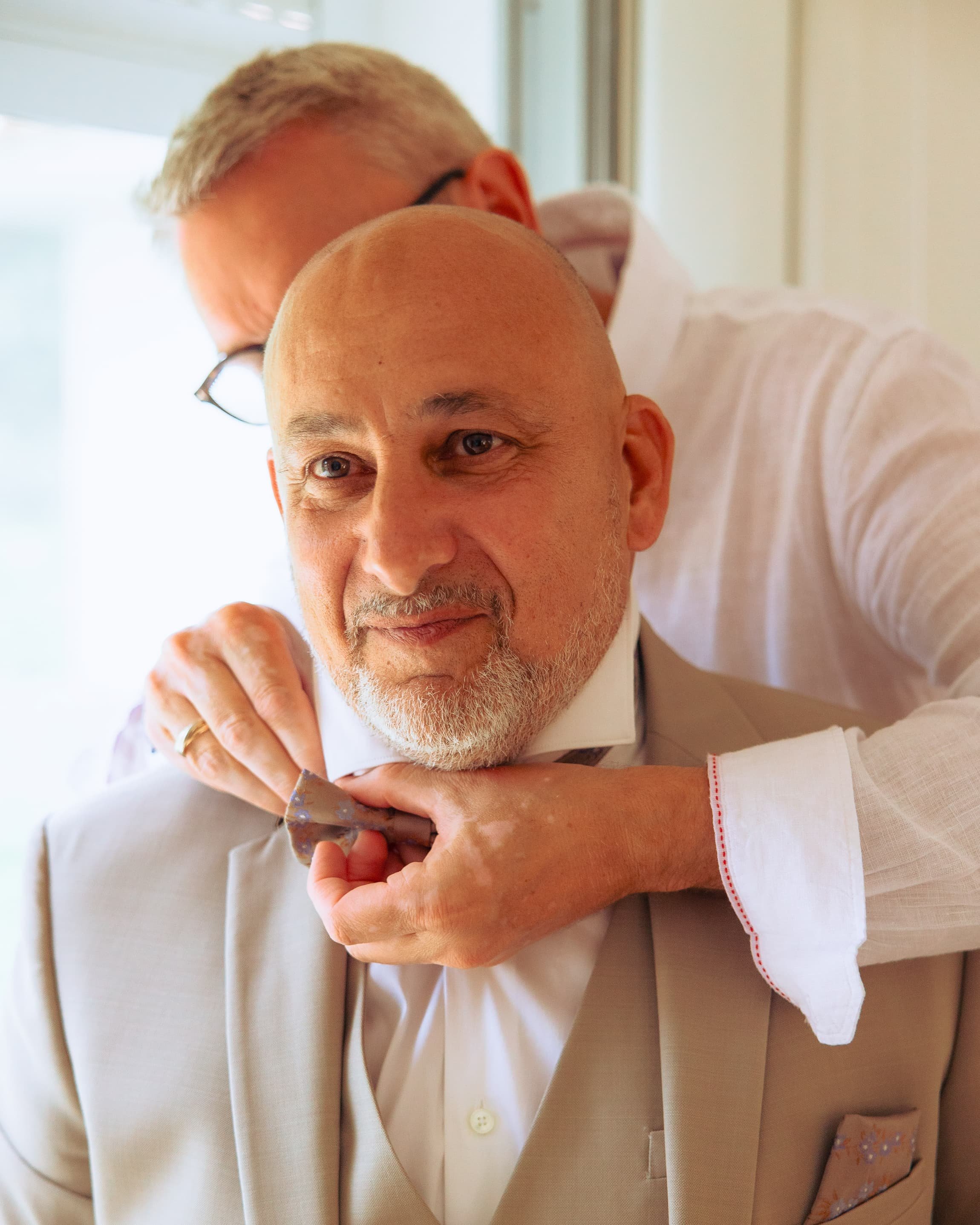 Deux hommes en costume blanc homme un dégusteur à un col blanc, l'autre en arrière-plan ajuste la cravate du premier dans un intérieur lumineux.