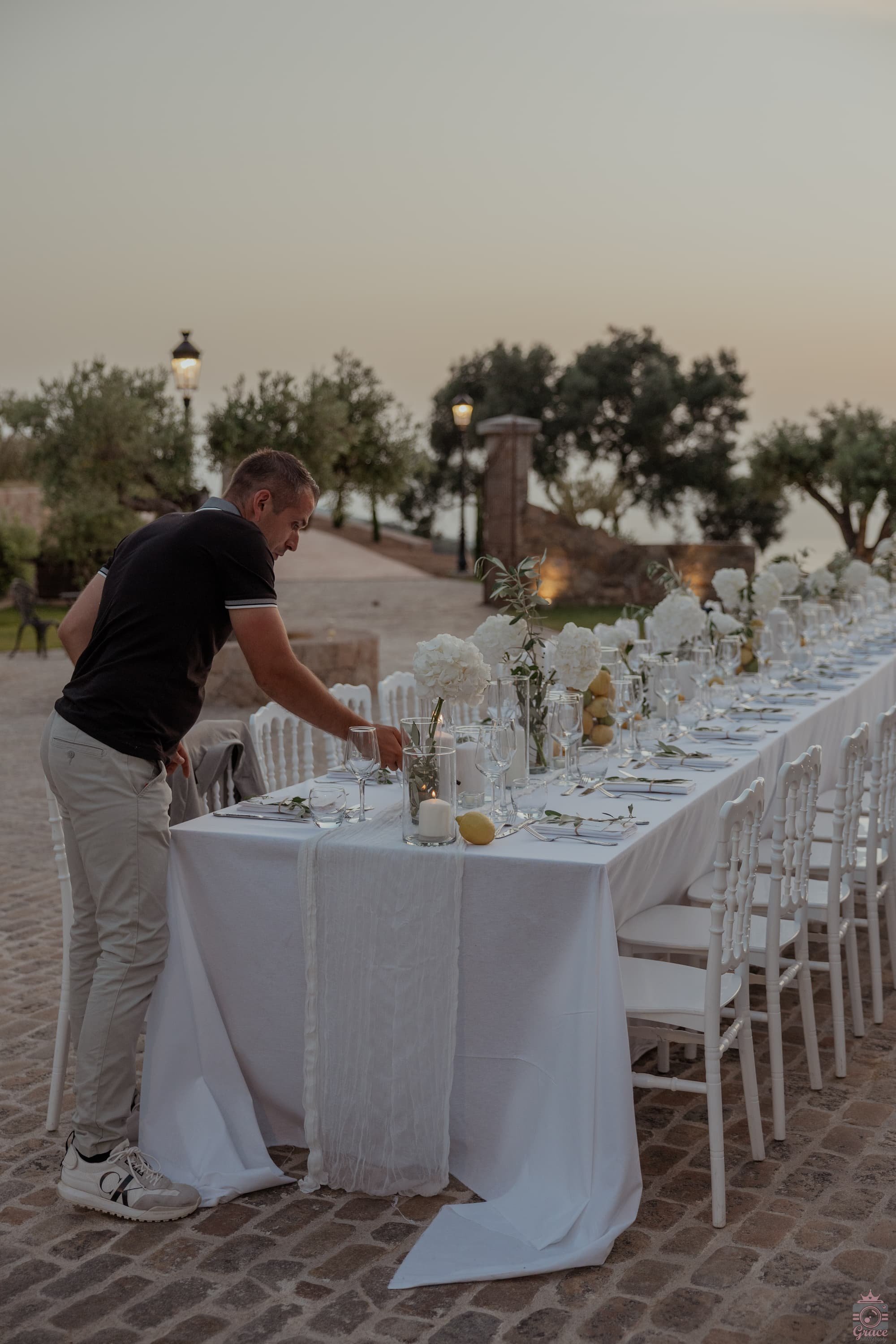 Homme préparant une table de dîner en plein air, décorée avec des fleurs blanches et des bougies, au crépuscule.